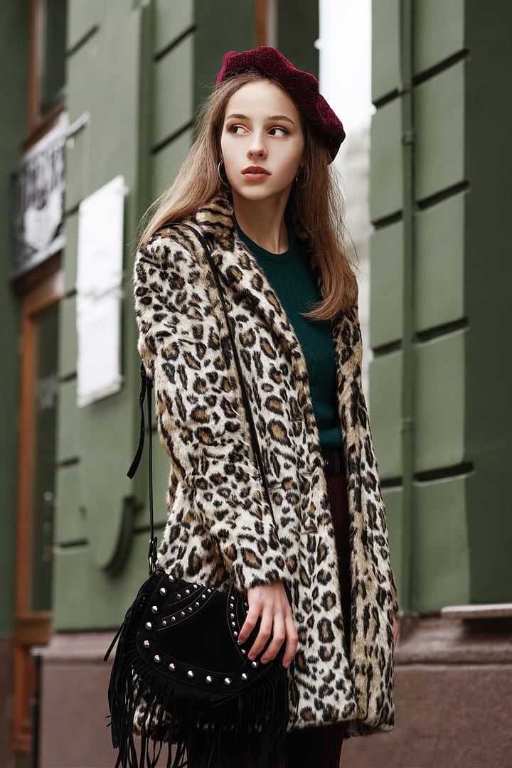 Outdoor fashion portrait of young beautiful fashionable lady wearing trendy animal, leopard print faux fur coat, beret, carrying suede shoulder bag with fringe, posing in street of European city