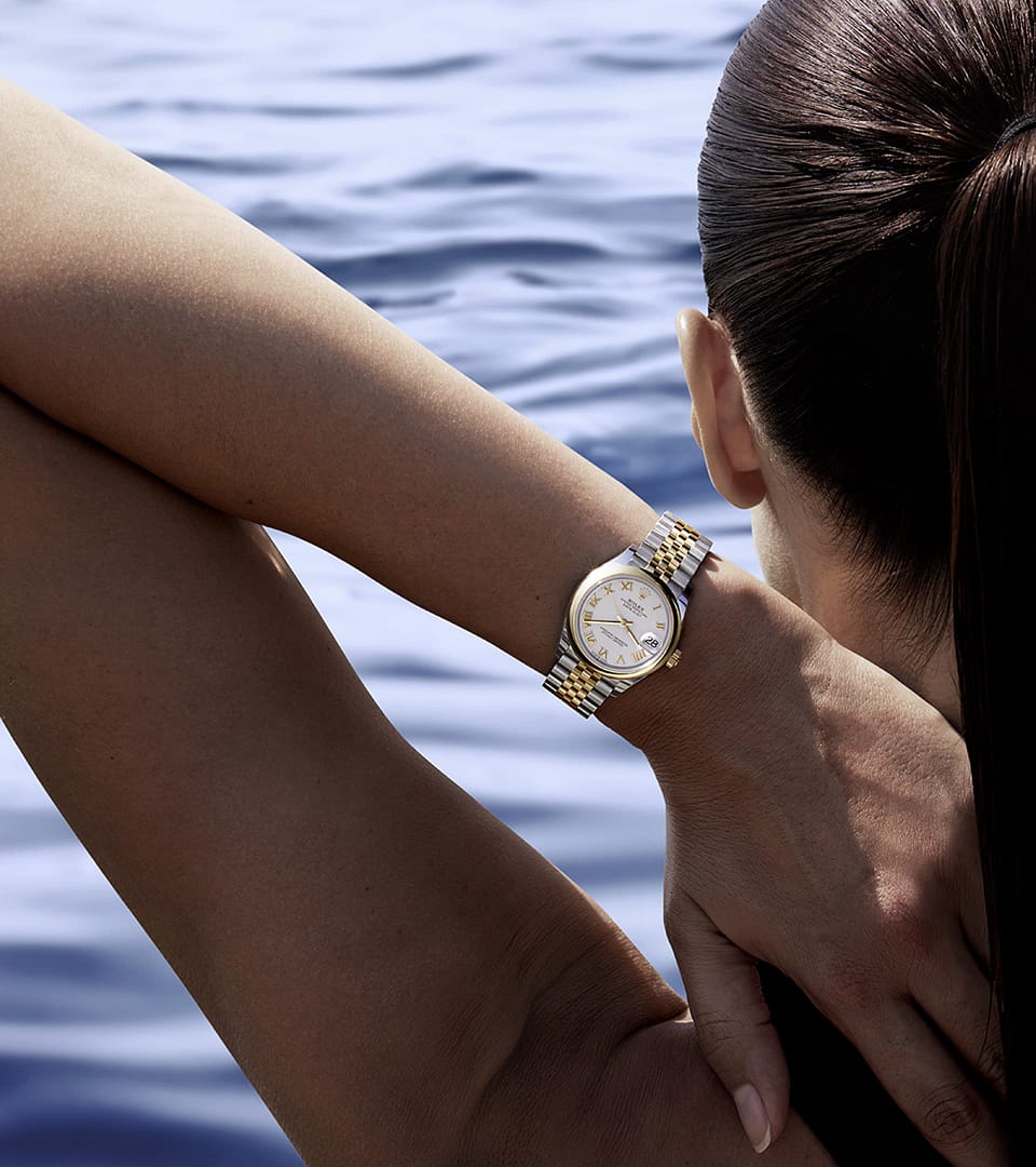Close-up of a person with long hair, showcasing a gleaming gold and silver Rolex Lady-Datejust wristwatch as they lean against their arm, with a serene body of water in the background