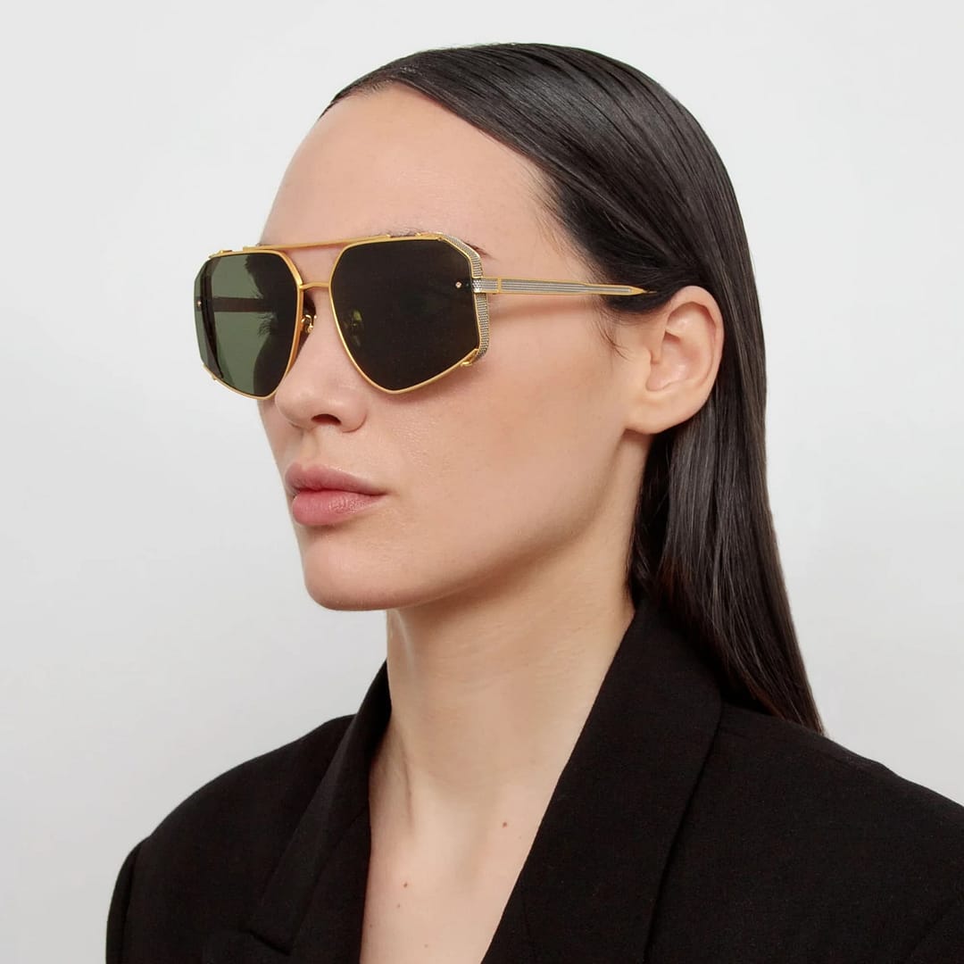 Woman with long dark hair wears gold-rimmed sunglasses, perfect for square faces, and a black blazer against a plain background