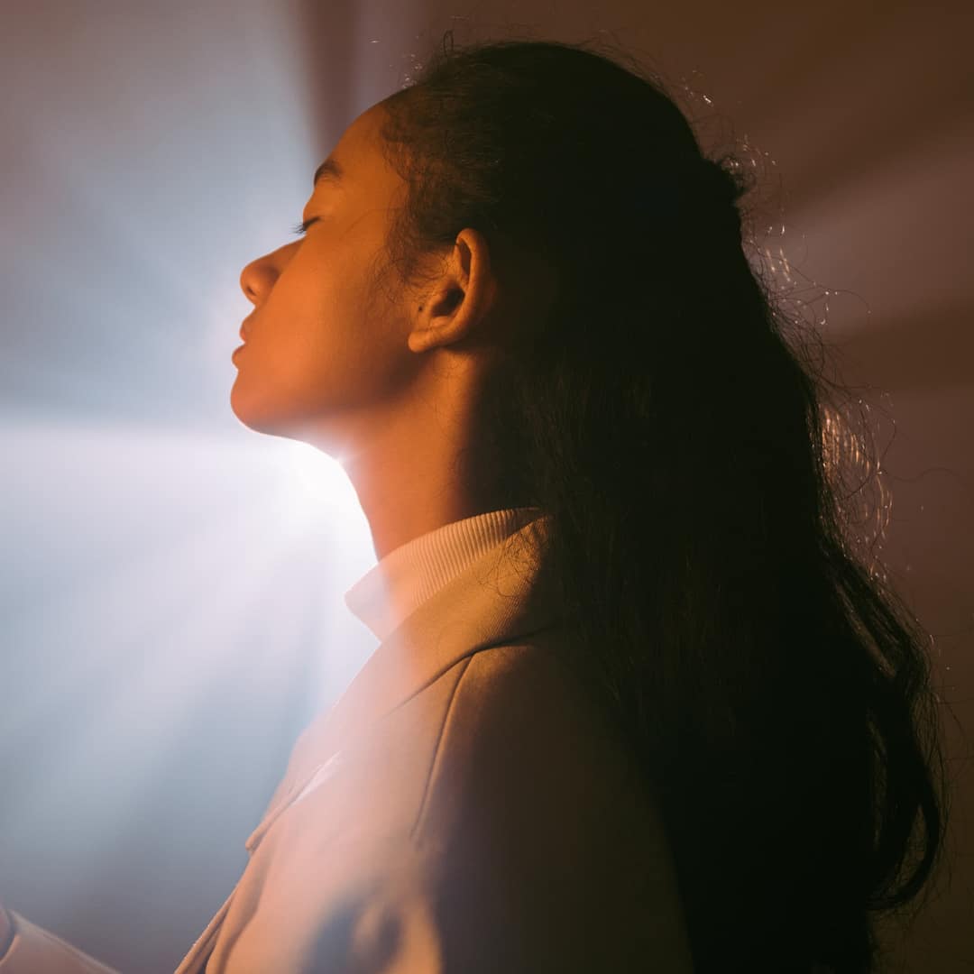 Woman in profile stands in front of a bright light source, creating a glowing, radiating effect behind her head that hints at self-love amid the dimly lit setting