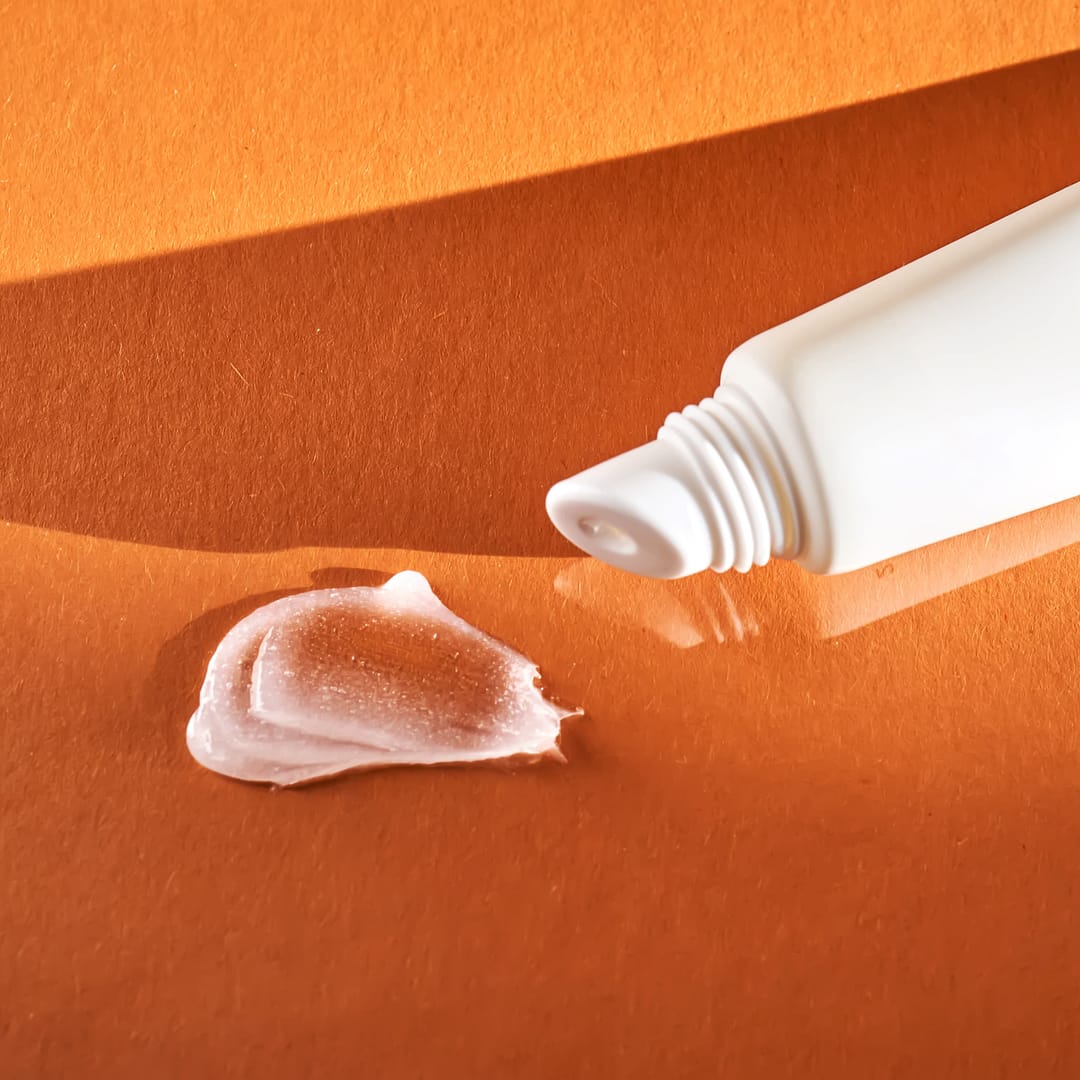A white squeeze tube of acne cream rests on an orange surface, its shadows softly cast alongside the delicate swirl of cream