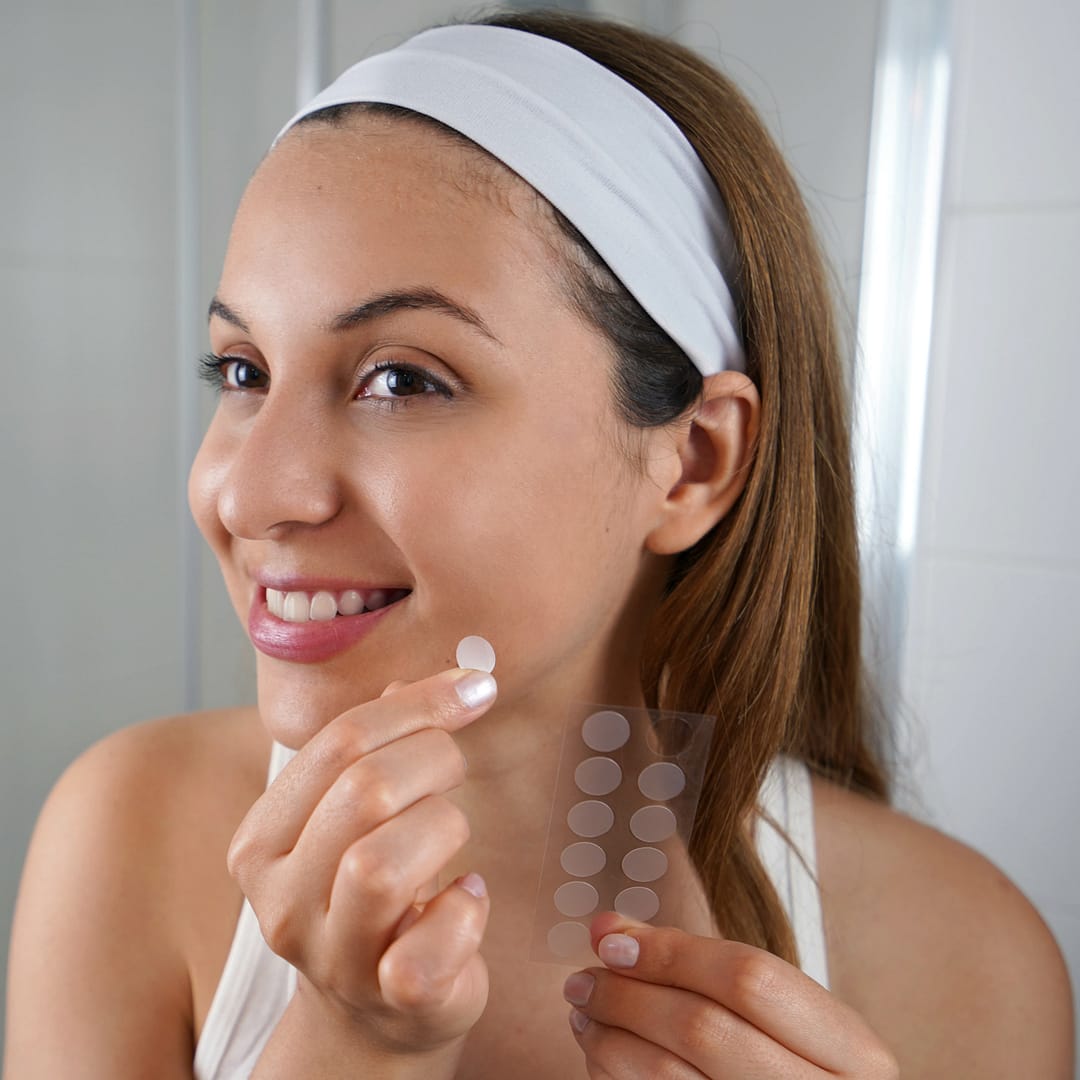 A woman with a headband smoothly applies a round, clear pimple patch from a sheet to her cheek