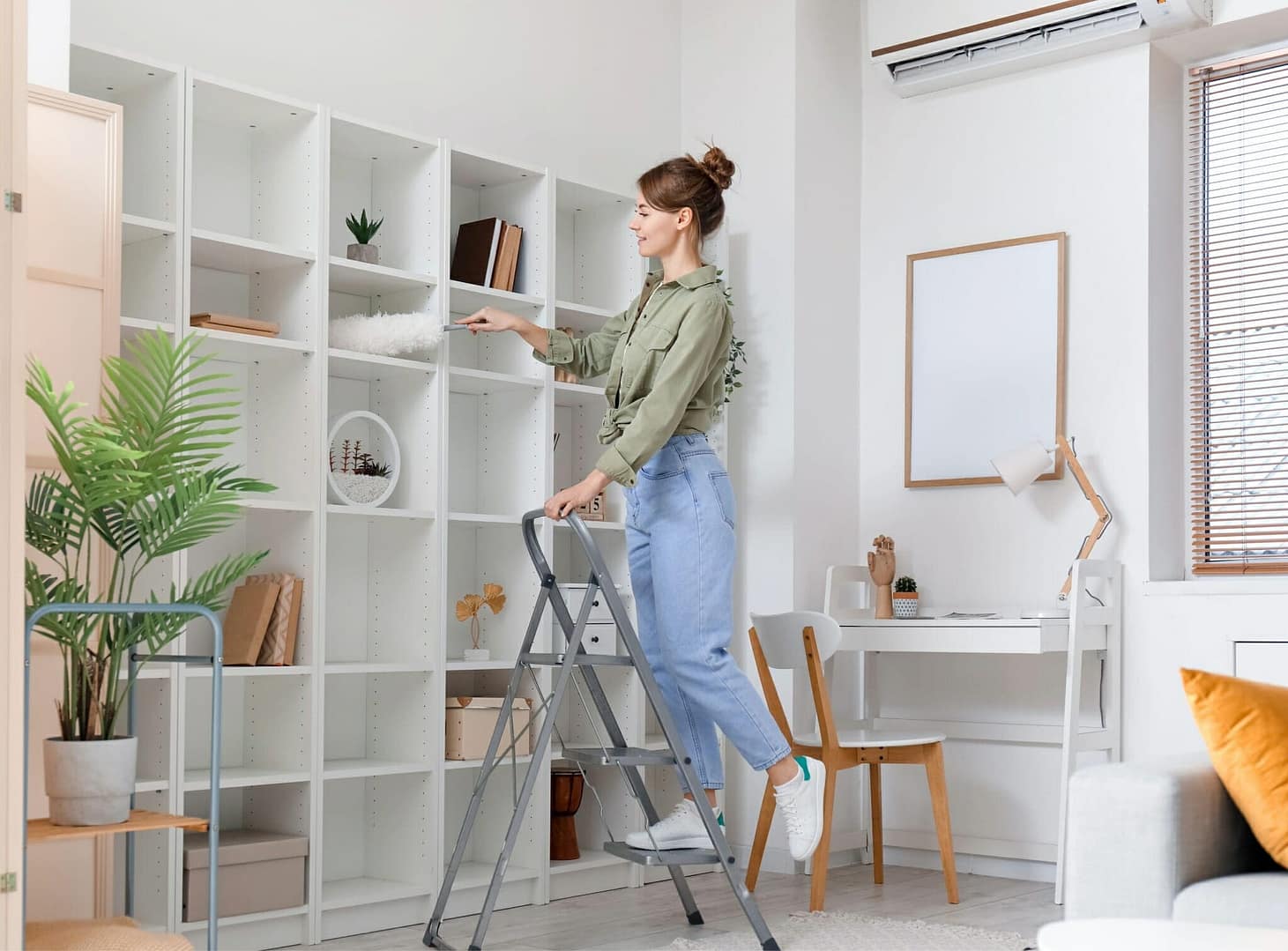 Young woman on stepladder cleaning bookshelf with duster at home