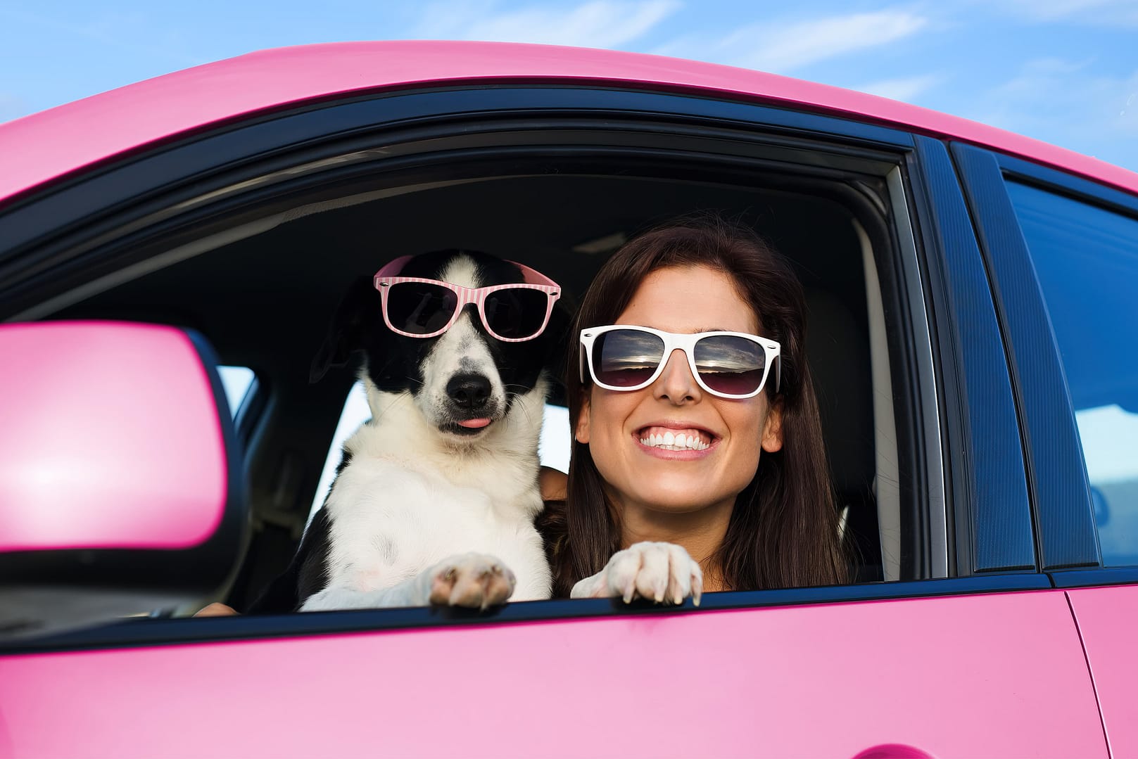 A woman and a dog wearing sunglasses, smiling from the window of a pink car