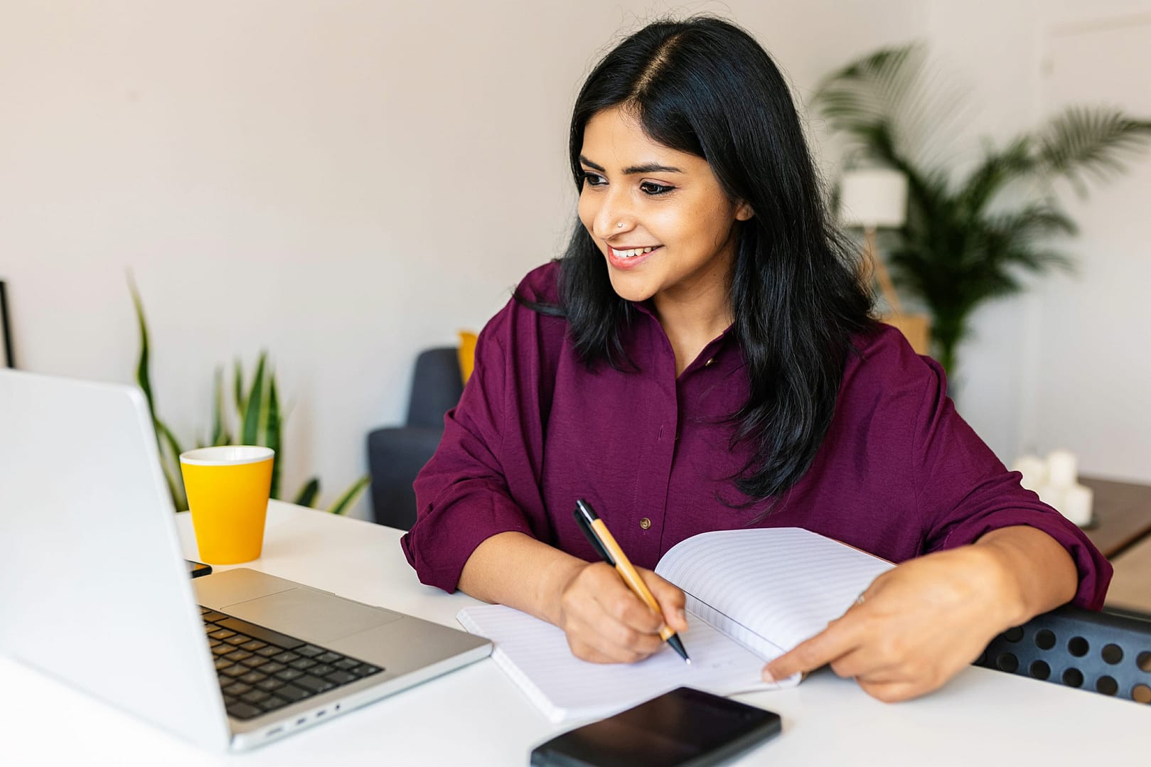 Young female student taking notes while using laptop computer at home