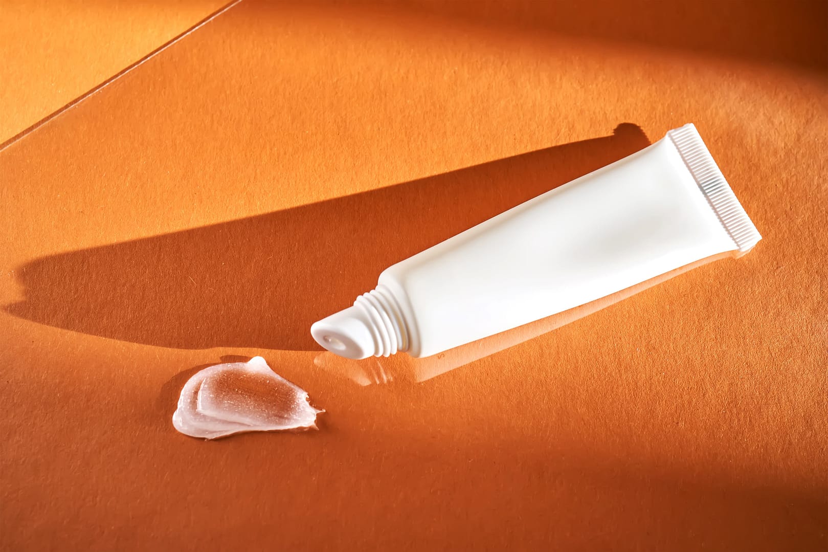 A white squeeze tube of acne cream rests on an orange surface, its shadows softly cast alongside the delicate swirl of cream