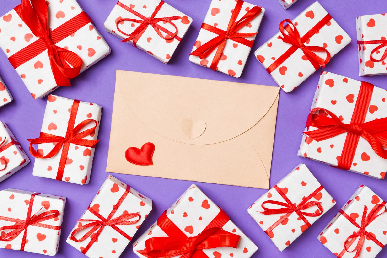 Several gift boxes wrapped in heart-patterned paper with red ribbons surround a beige envelope featuring a small heart sticker, placed on a purple background