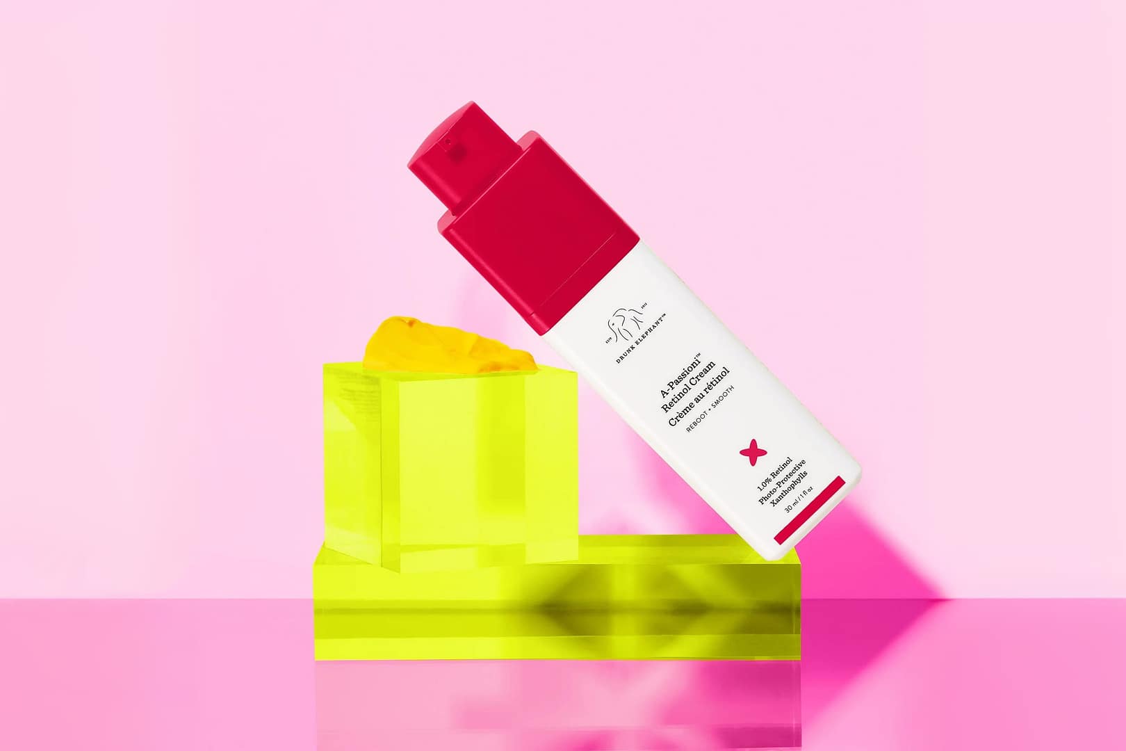 The vibrant bottle of Drunk Elephant A-Passioni Retinol Cream stands poised above a yellow geometric object, set against a charming pink backdrop