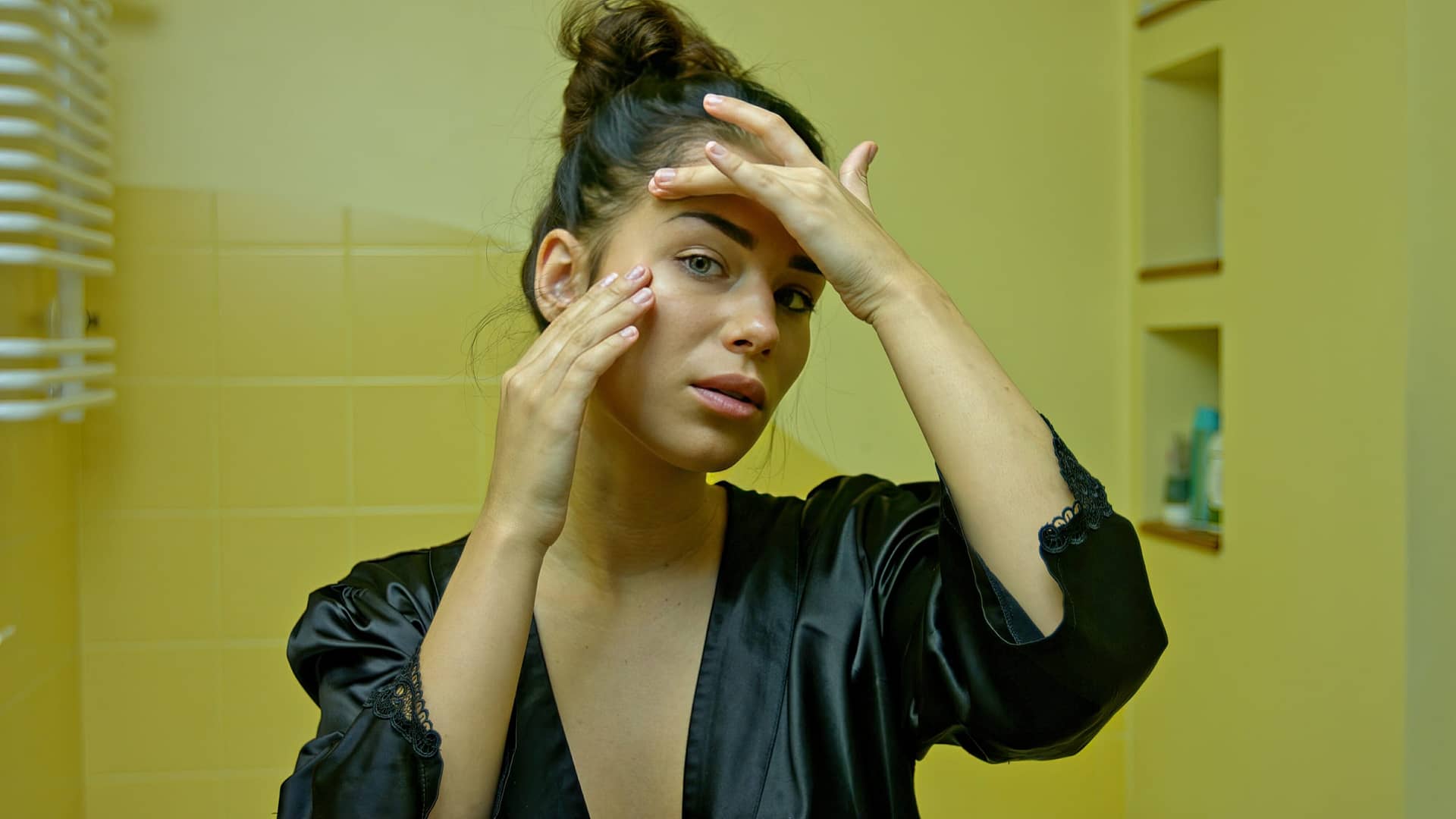 A young woman in the home bathroom looks in the mirror and checks her first wrinkles