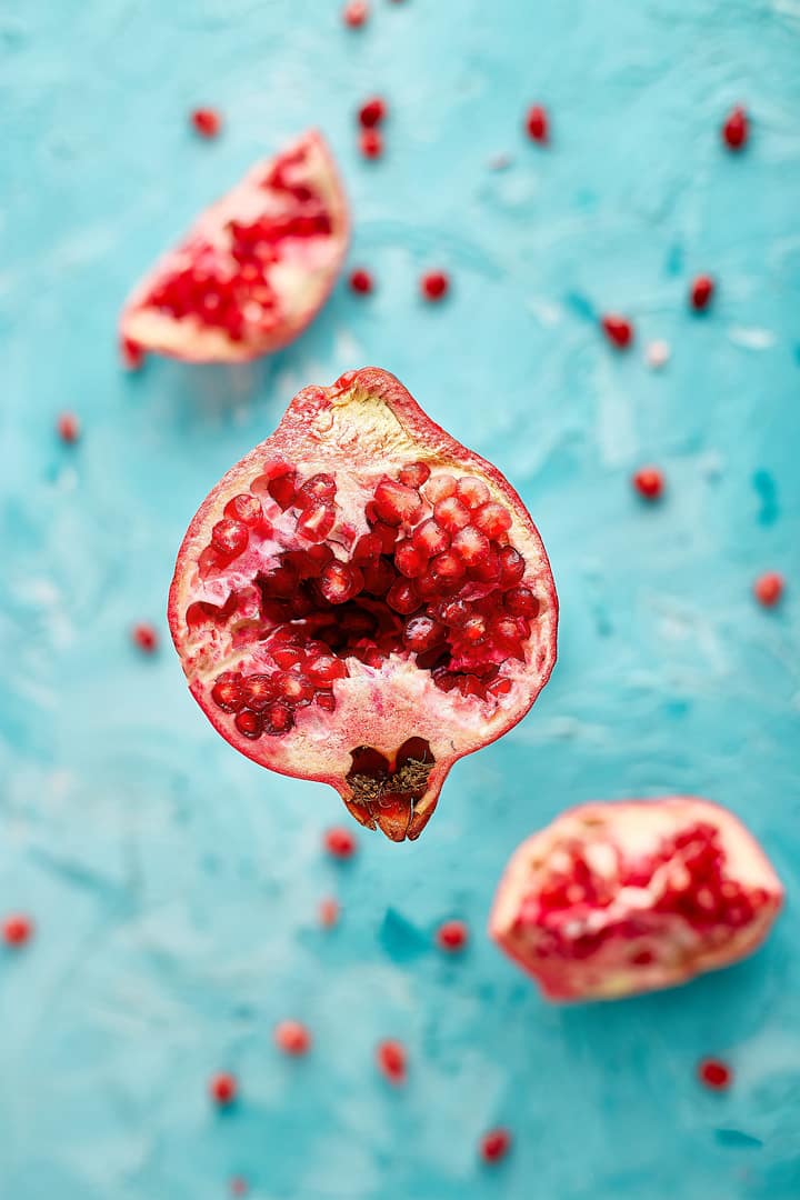 Pomegranate for glowing skin. A halved pomegranate with seeds exposed is surrounded by scattered seeds and additional pomegranate pieces against a textured blue background
