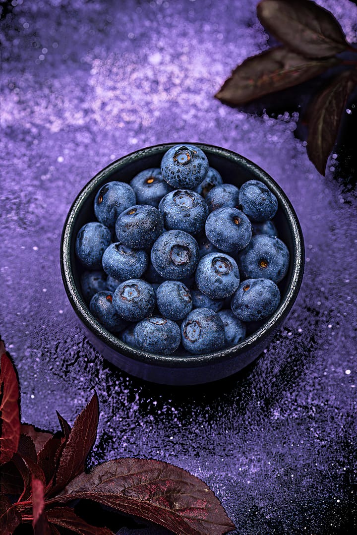 Blueberries for glowing skin. A black bowl filled with fresh blueberries sits on a purple textured surface, surrounded by dark red leaves