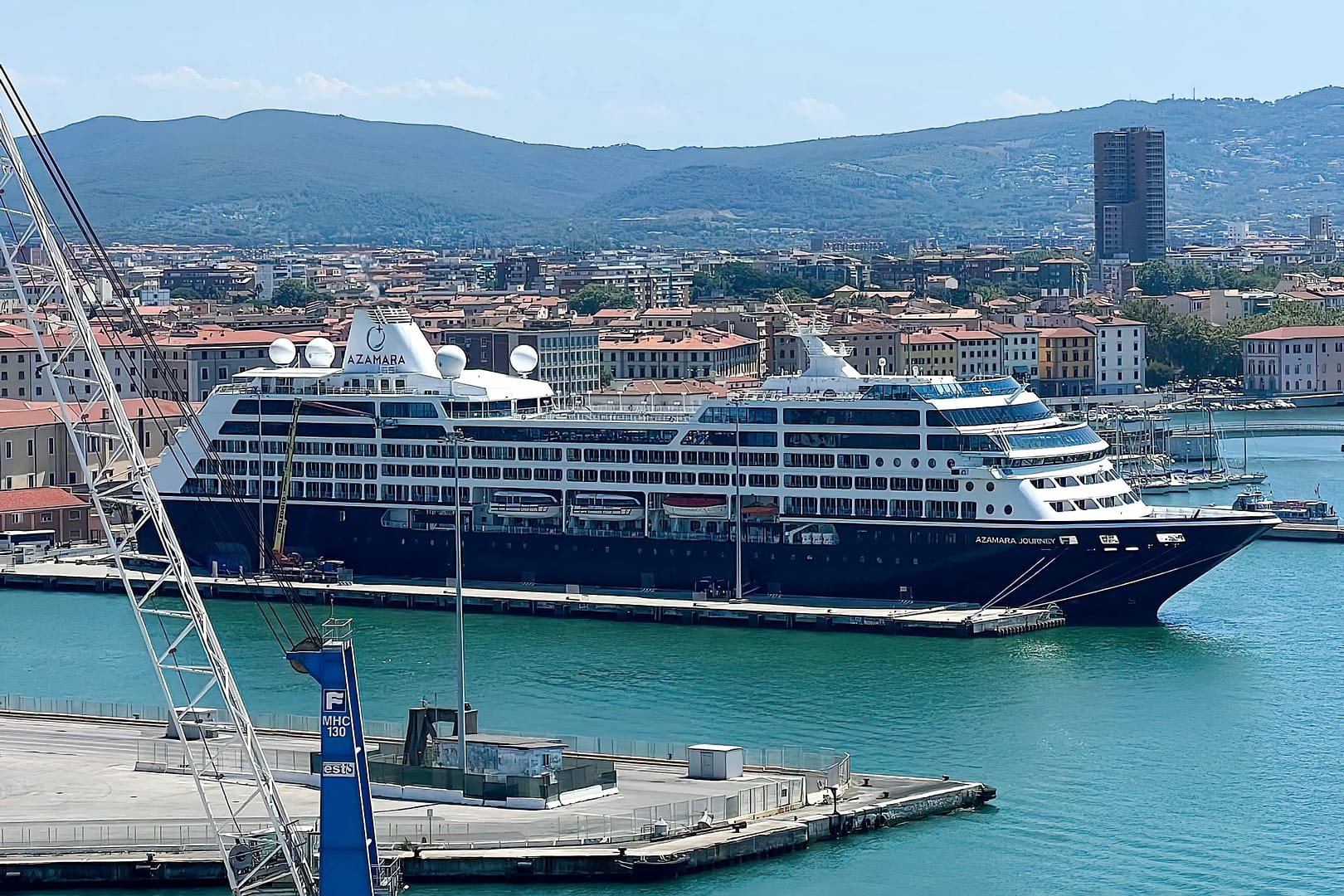 A large Azamara Journey cruise ship docked at a pier in a coastal city with hills in the background and clear blue skies overhead