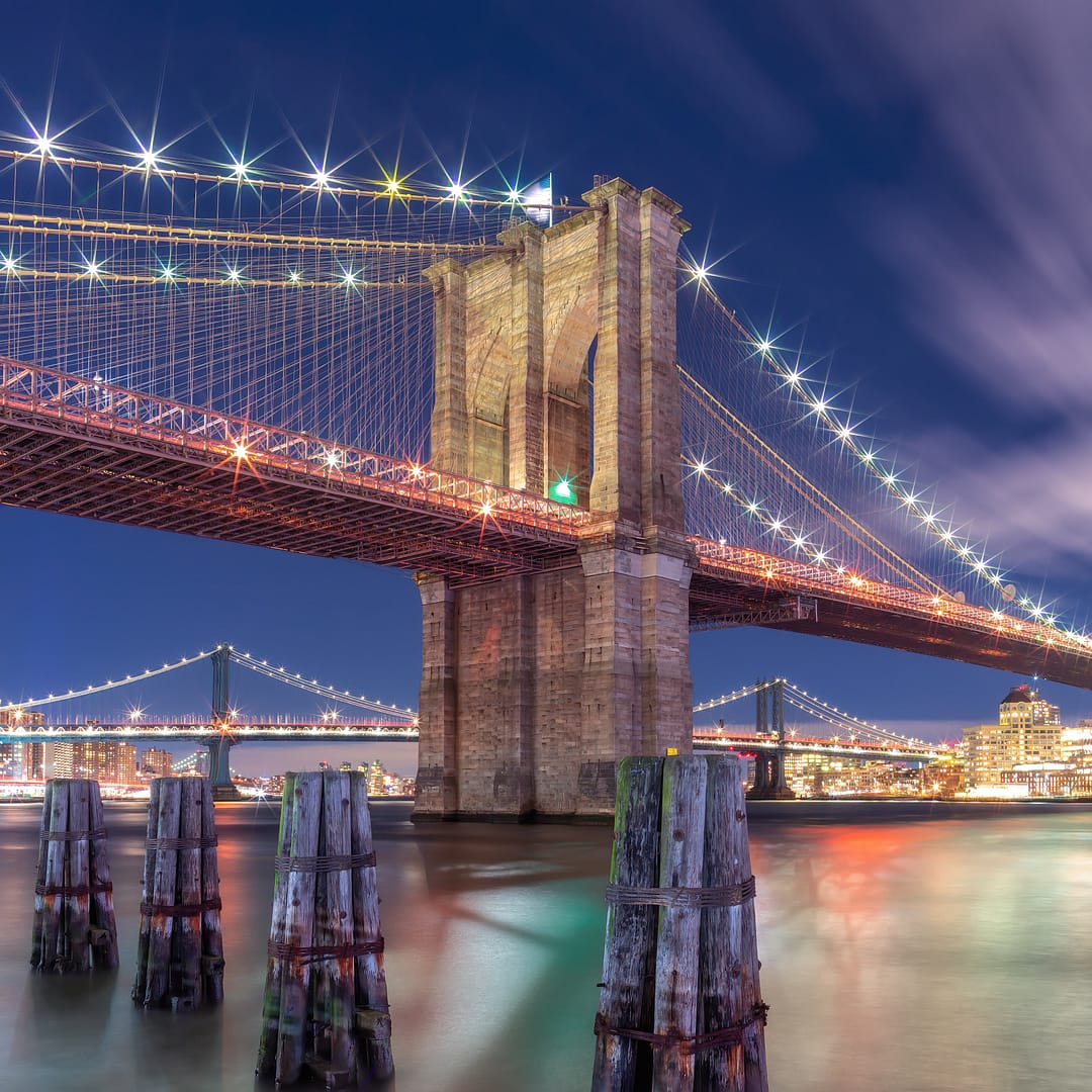 Nighttime view of the Brooklyn Bridge illuminated, with smooth water and skyline in the background