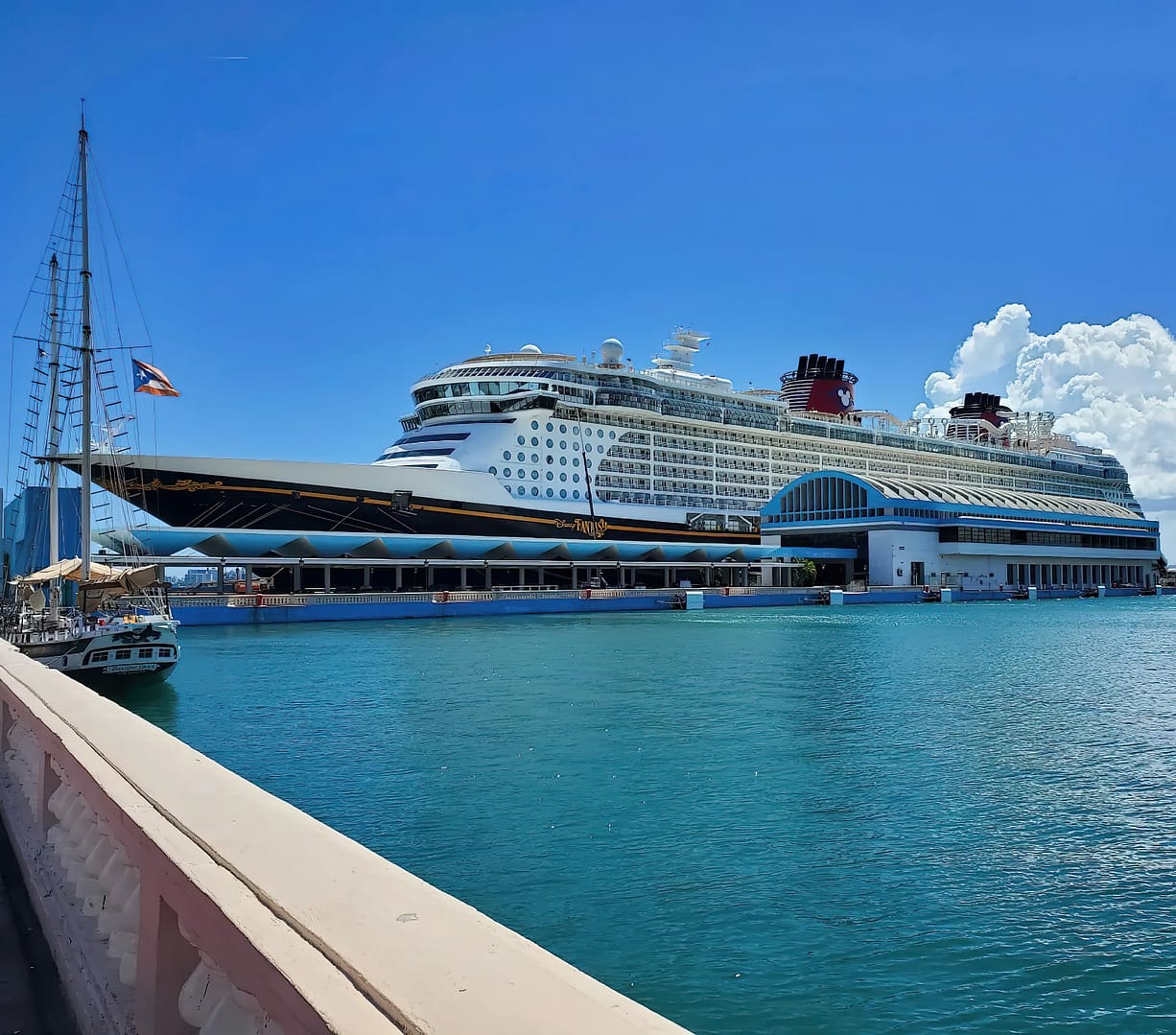 A large Disney Fantasy cruise ship docked at a port with a clear blue sky and calm sea waters in the foreground