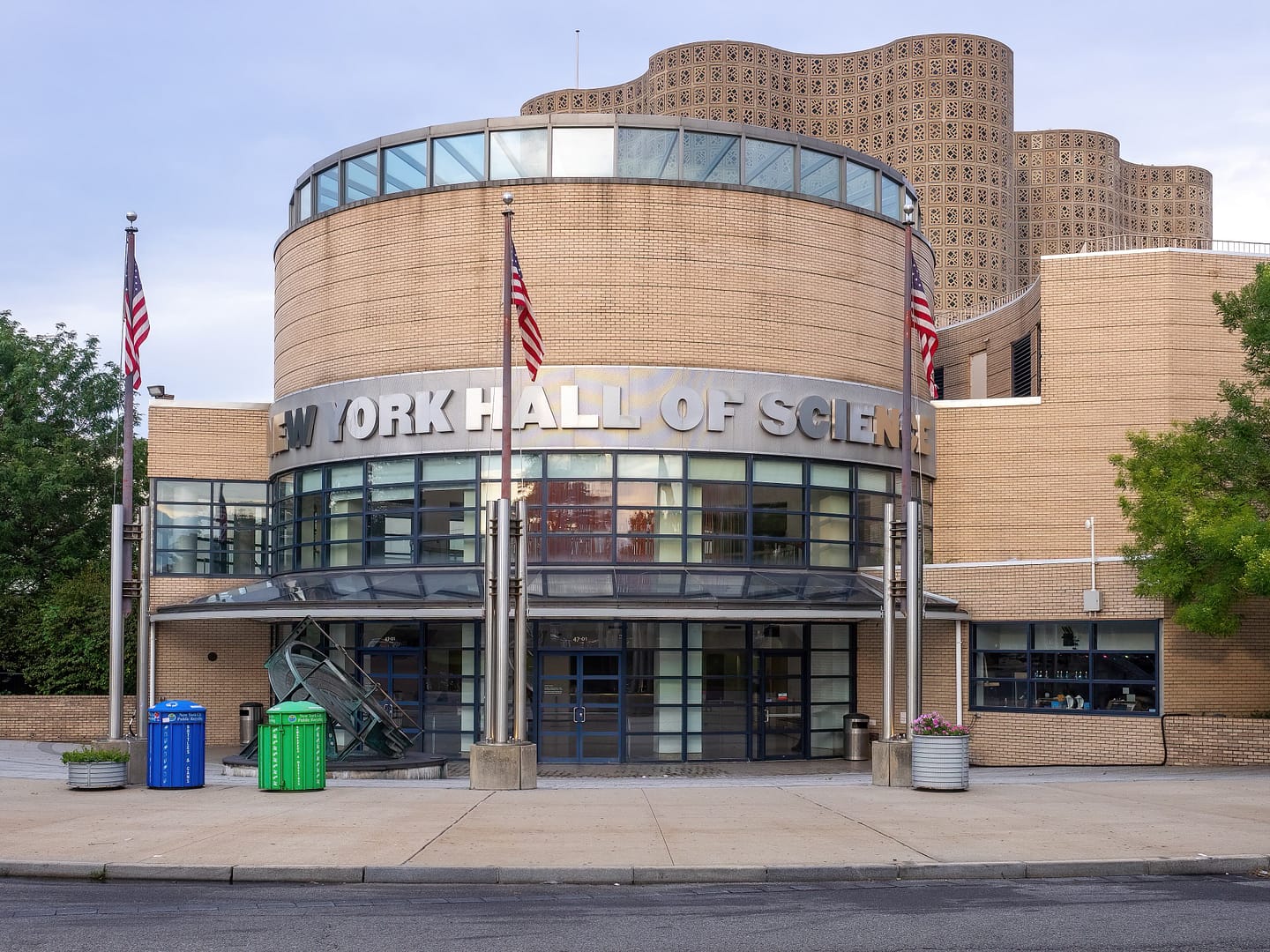 Front view of the New York Hall of Science building with a curved design, flanked by two American flags, under a cloudy sky