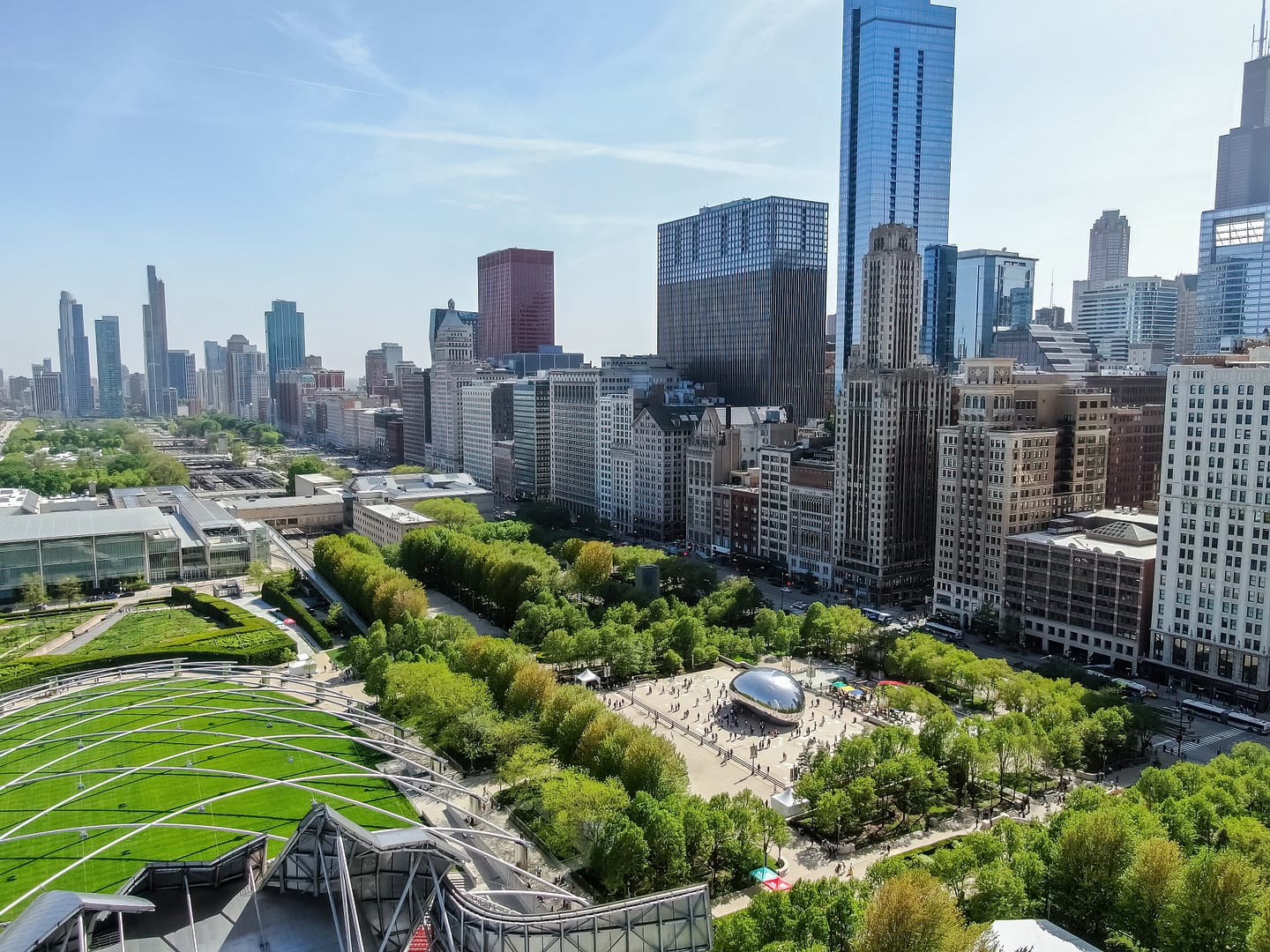 Aerial view of Millennium Park with a shiny, bean-shaped sculpture, surrounded by lush greenery and modern skyscrapers under a clear blue sky