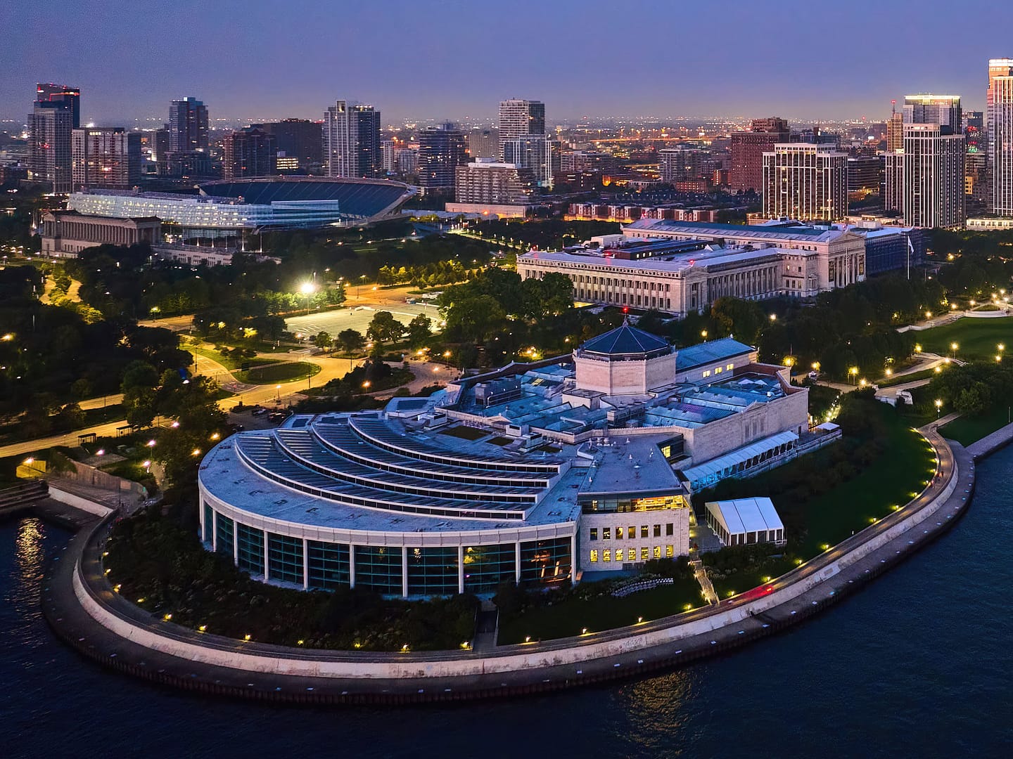 Aerial view of the modern, circular Shedd Aquarium by the waterfront at twilight, surrounded by a cityscape with illuminated buildings