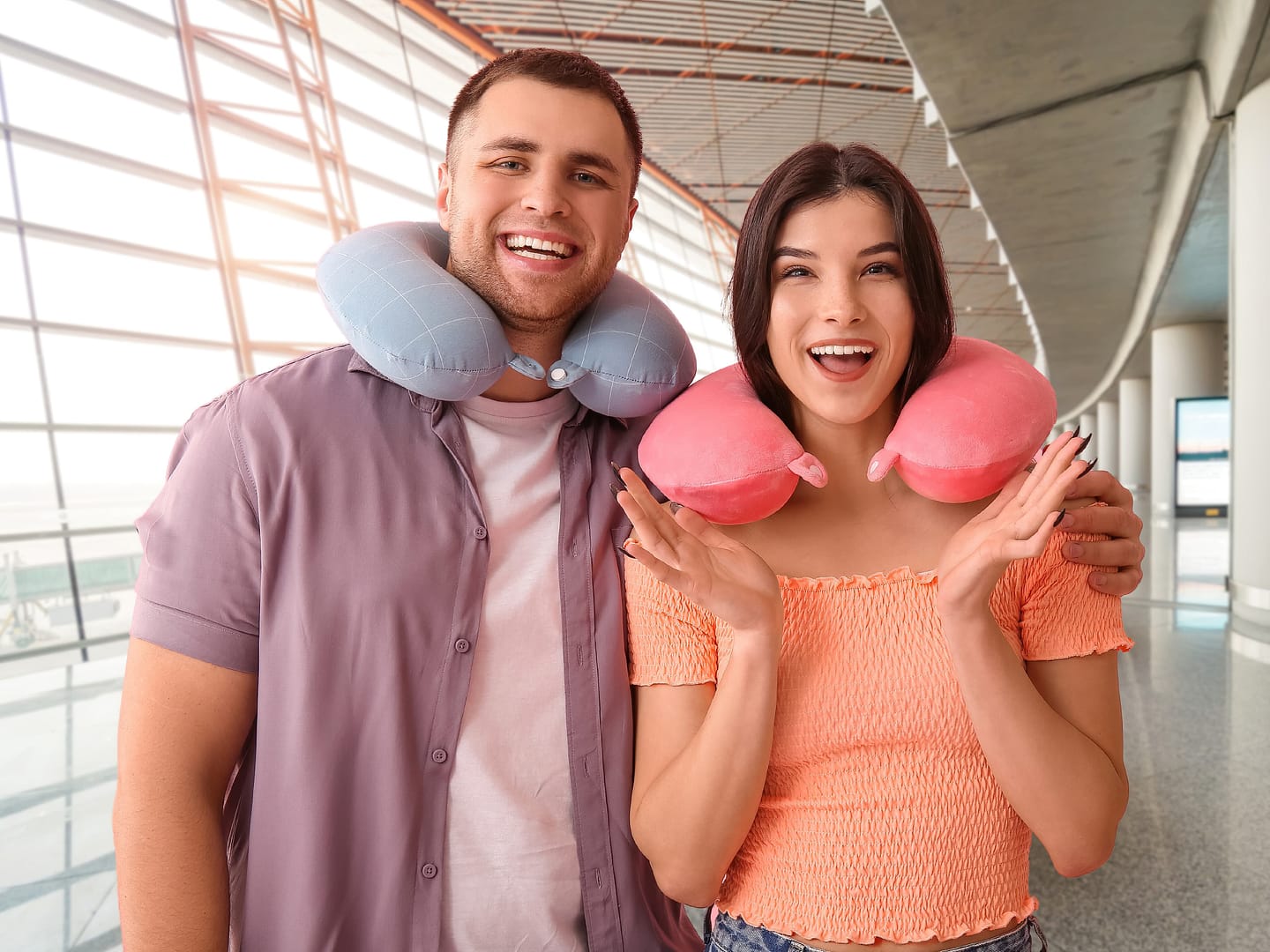 A cheerful young couple with travel neck pillows on their neck in an airport, smiling excitedly at the camera