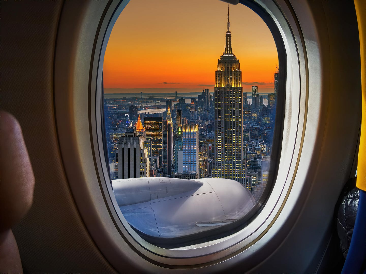 View of the New York City skyline at sunset seen through an airplane window, highlighting illuminated Empire State Building and other buildings in the center. Partial view of the airplane wing is visible