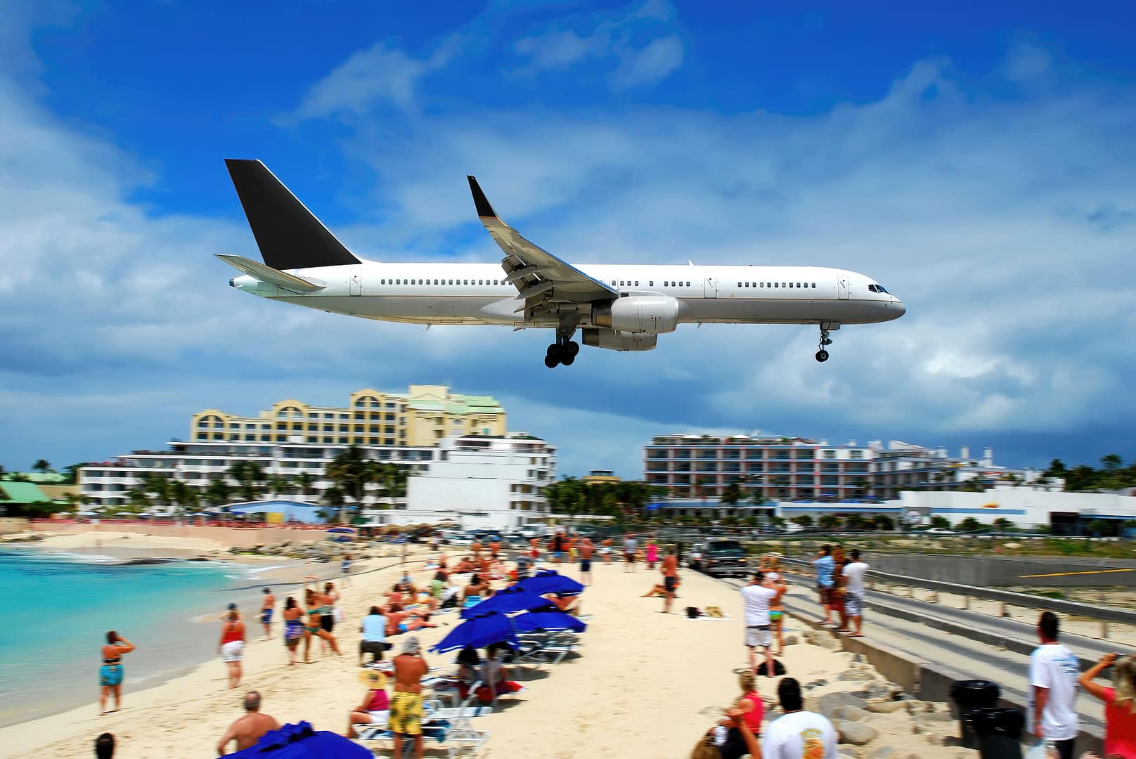 A commercial airplane landing low over a crowded beach near coastal buildings in St. Maarten on a sunny day