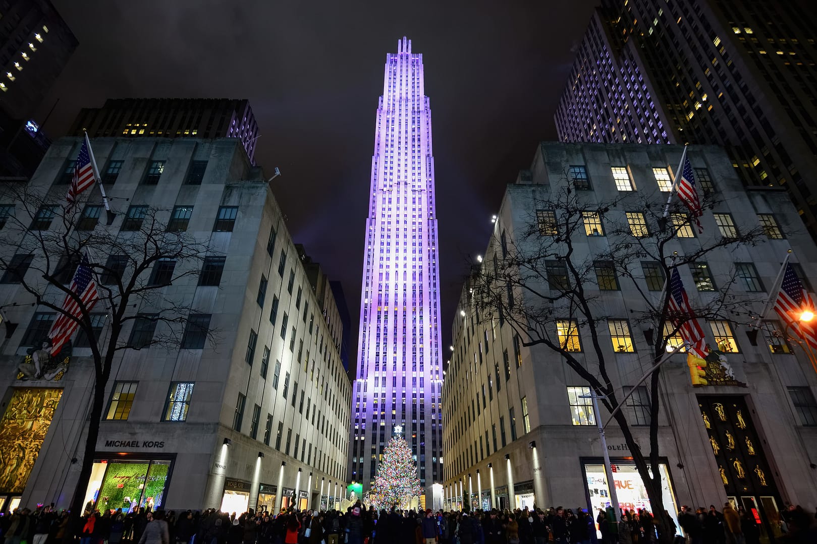 The Rockefeller Center in New York City illuminated in purple at night, showcasing the towering skyscraper and surrounding buildings, with a large, lit Christmas tree at its base