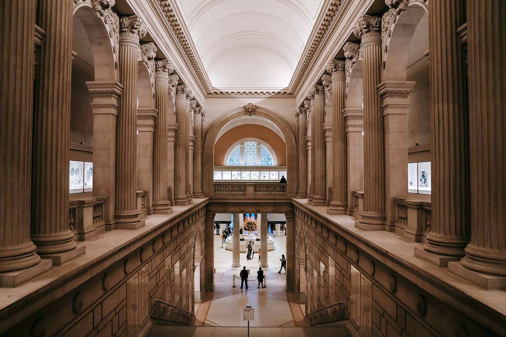Interior view of The Metropolitan Museum of Art hall with high arches and columns, featuring visitors walking and viewing exhibits, and a brightly lit stained glass window at the far end