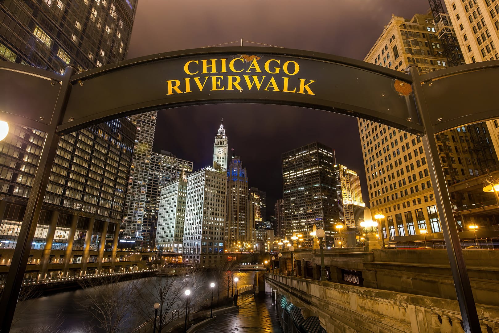 Night view of the Chicago Riverwalk sign with illuminated skyscrapers and the Wrigley Building in the background