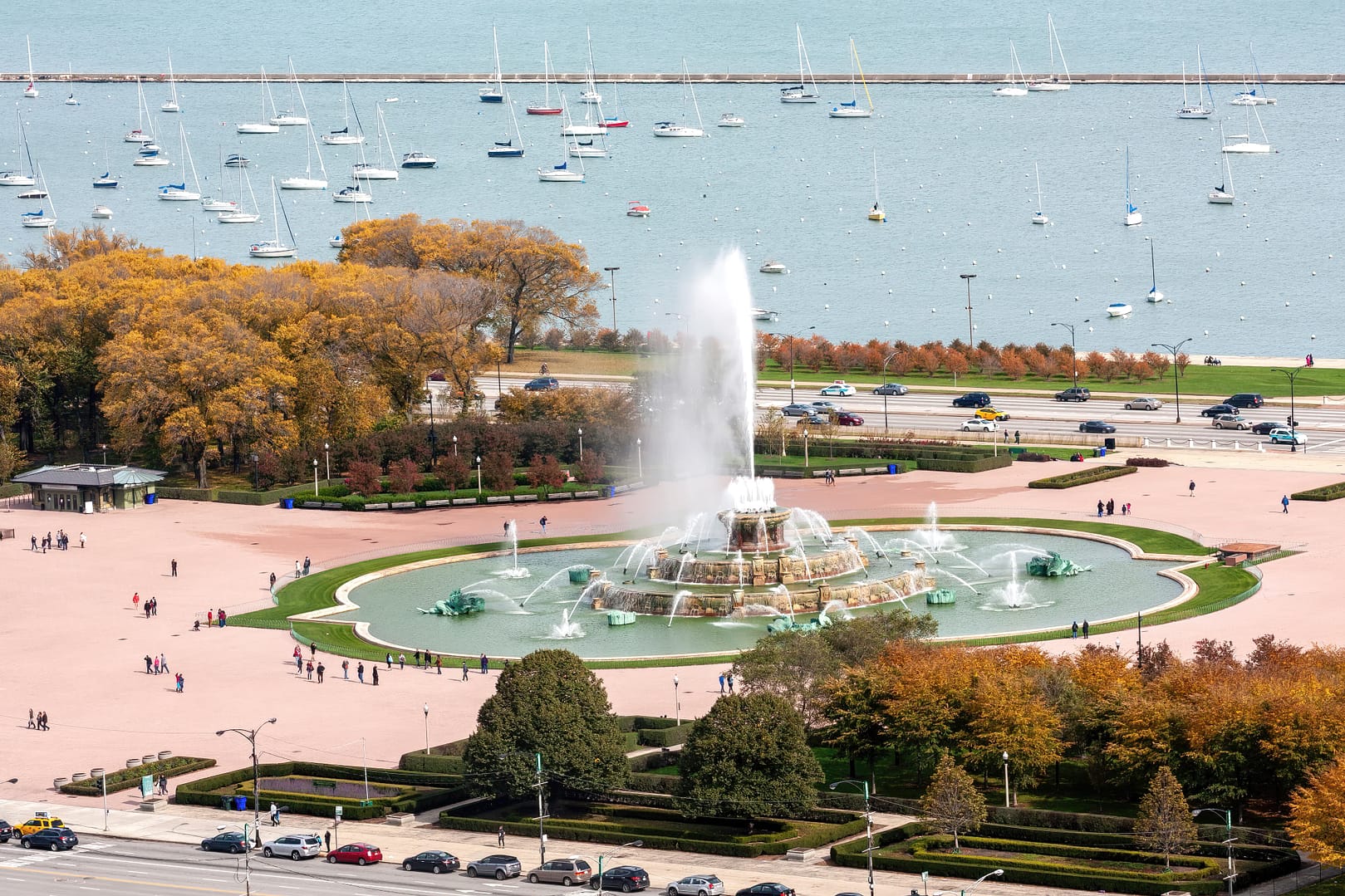 Aerial view of Buckingham Fountain in Grant Park, Chicago, operating with a large water spray, surrounded by trees and overlooking a marina with sailboats