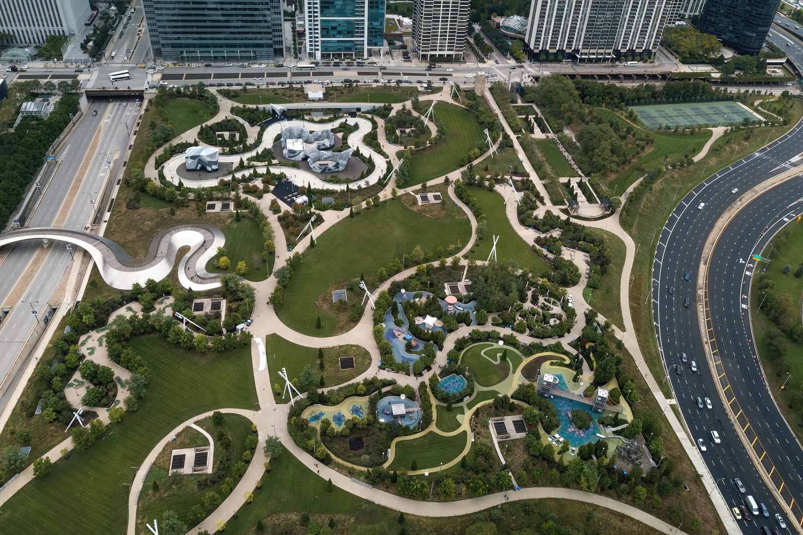 Aerial view of Maggie Daley Park with its intricately designed layout featuring winding paths, water features, and lush greenery, adjacent to a busy highway