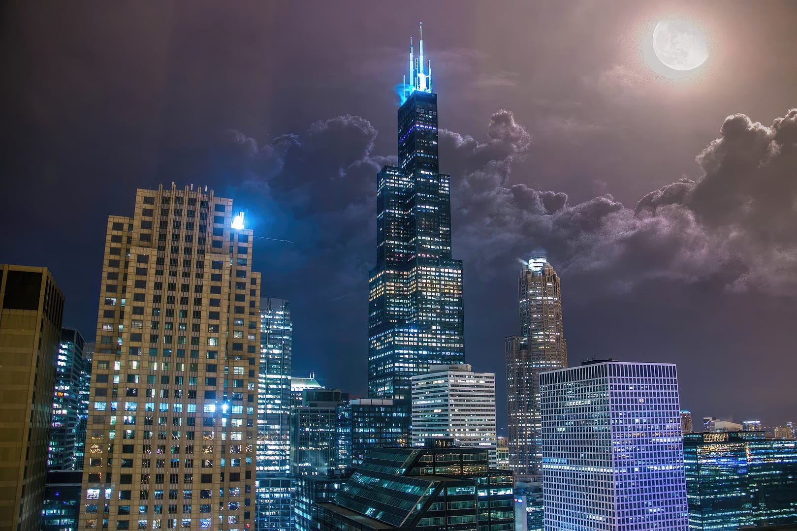 Nighttime cityscape showcasing the illuminated Willis Tower under a full moon with surrounding skyscrapers in Chicago