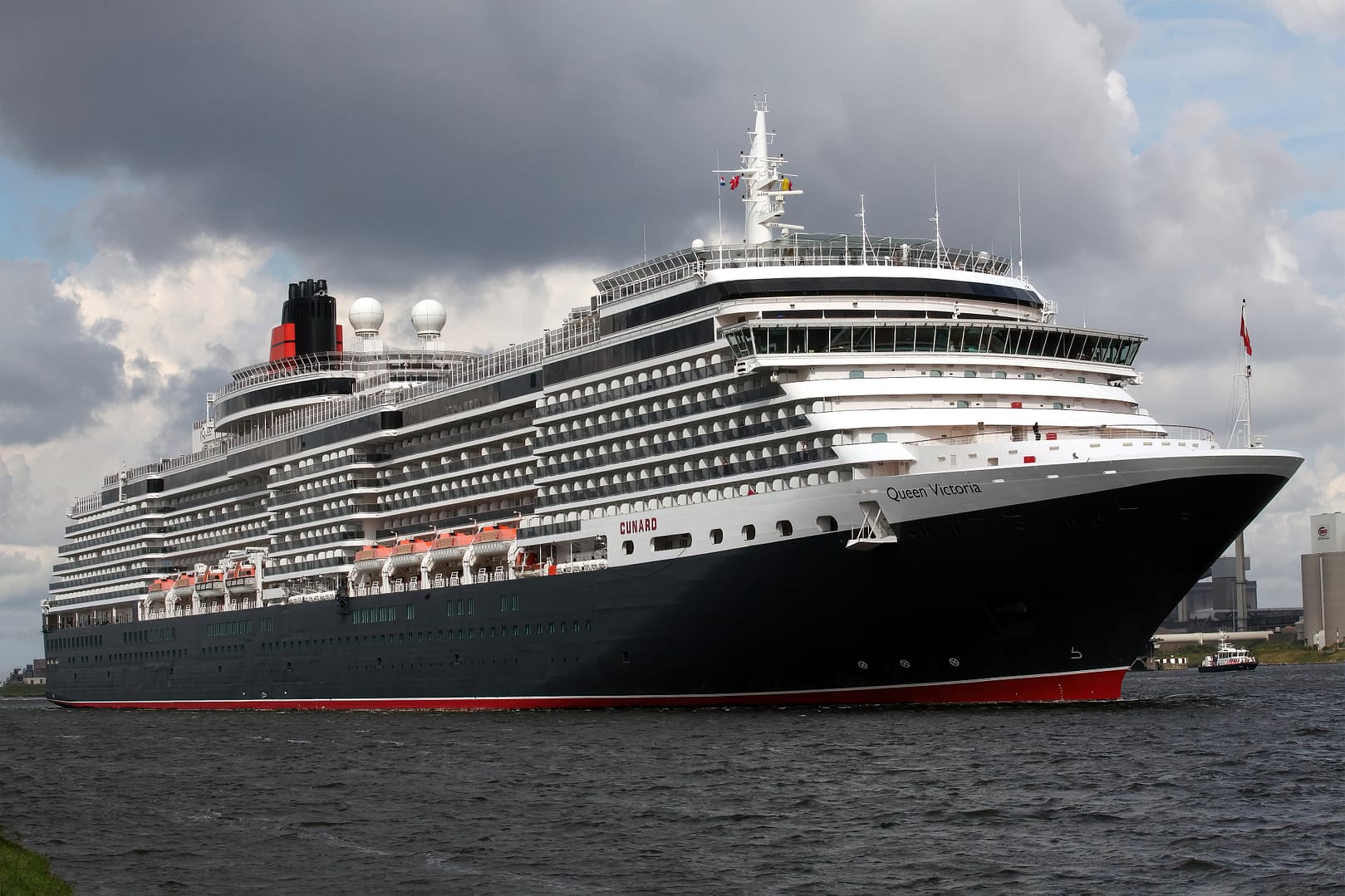 Large Cunard Queen Victoria cruise ship with black and red hull on water under cloudy sky, with multiple decks visible and smokestacks at the top