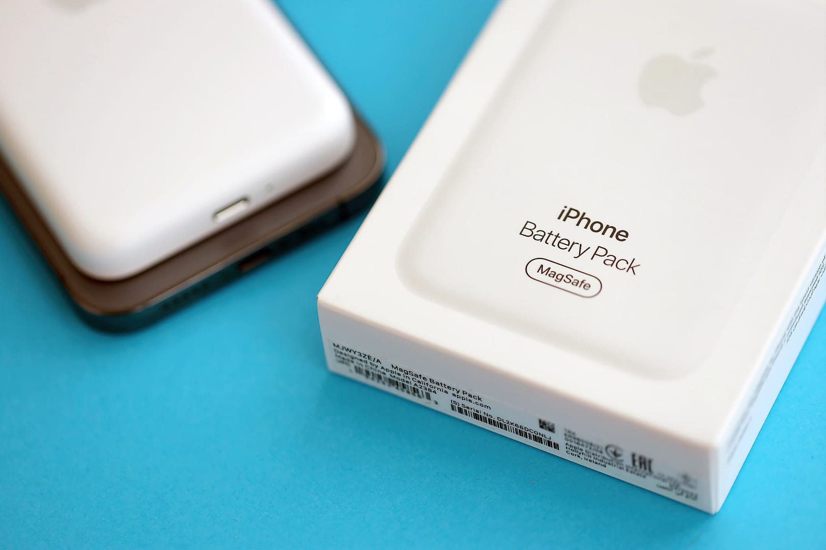 An iPhone's MagSafe Battery Pack in its packaging, with a blurred iPhone in the background, placed on a blue surface