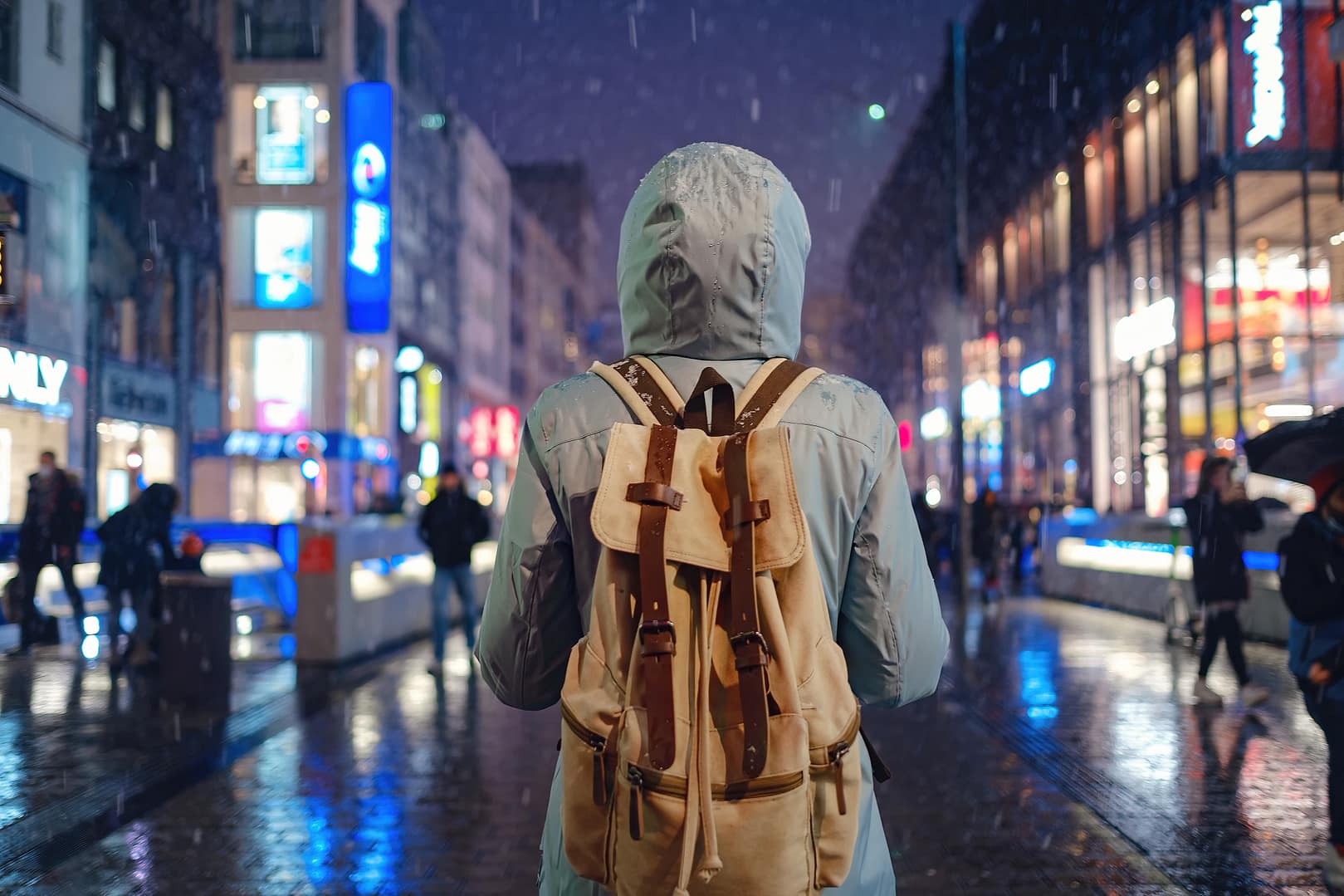 Traveler wearing a hooded jacket and backpack standing alone on a rainy city street at night, with illuminated shop signs and pedestrians in the background