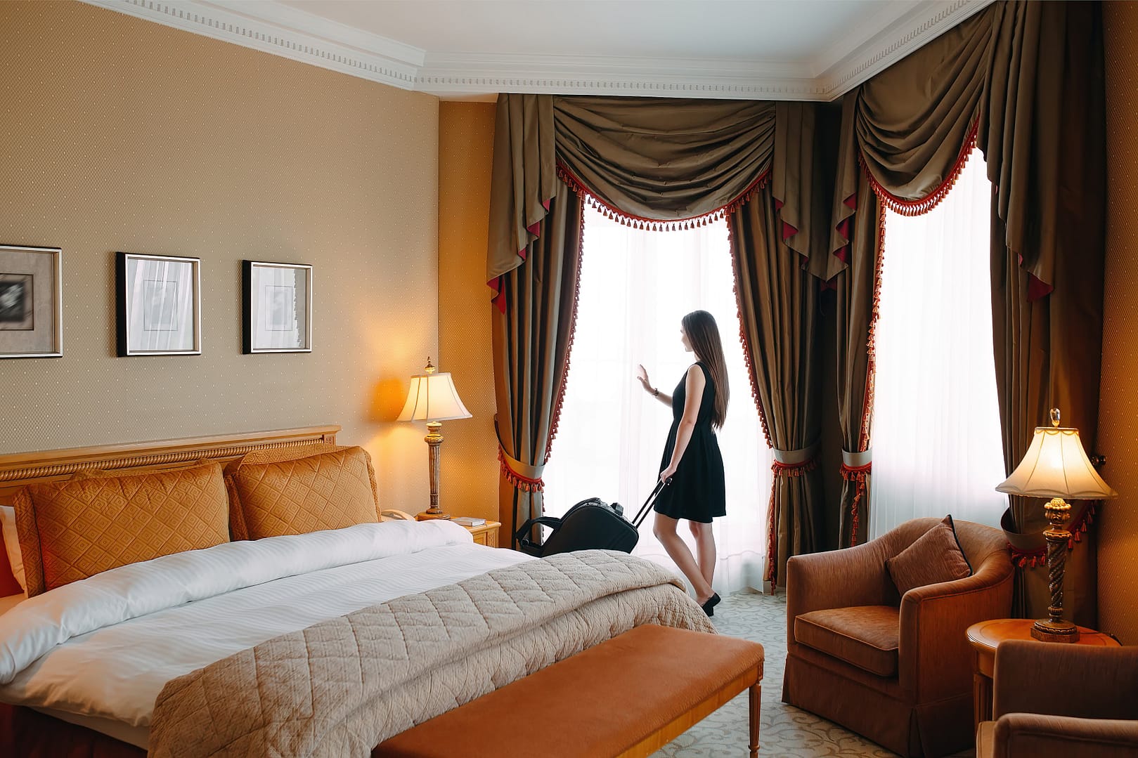 A woman in a little black dress standing by a window in a well-lit, luxurious hotel room, looking outside, with a suitcase nearby