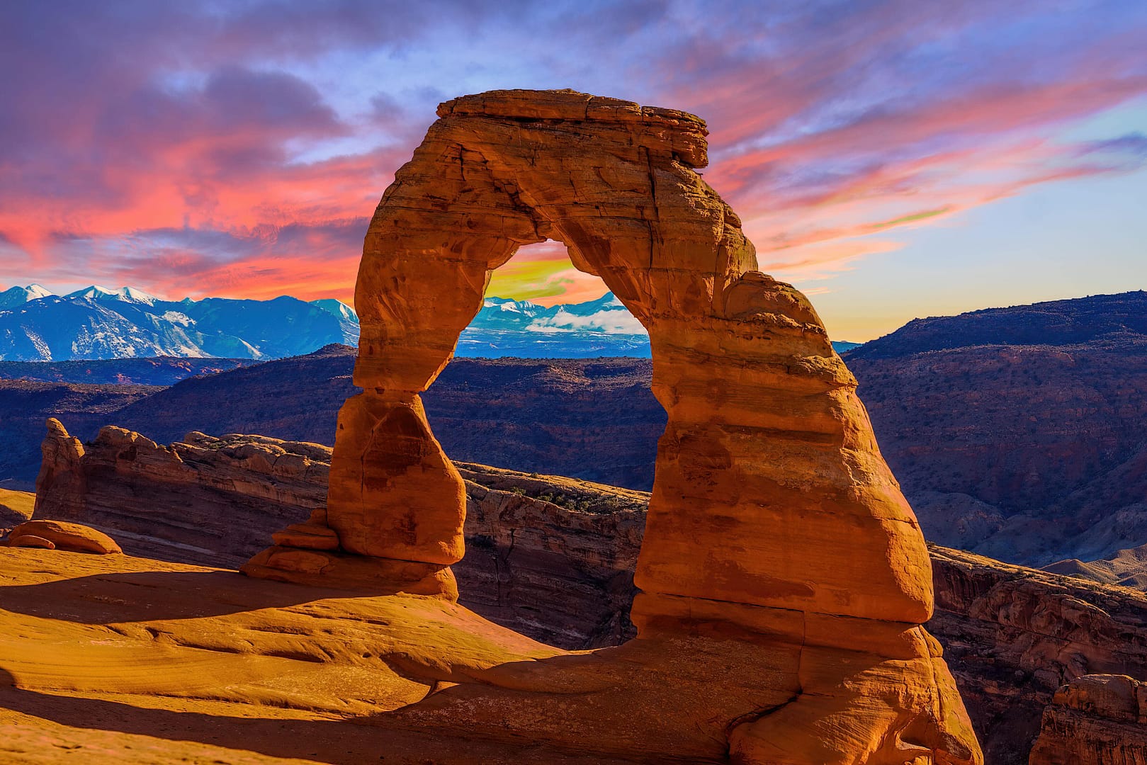 A natural sandstone arch stands against a backdrop of colorful sunset skies and distant mountains in Arches National Park in Utah