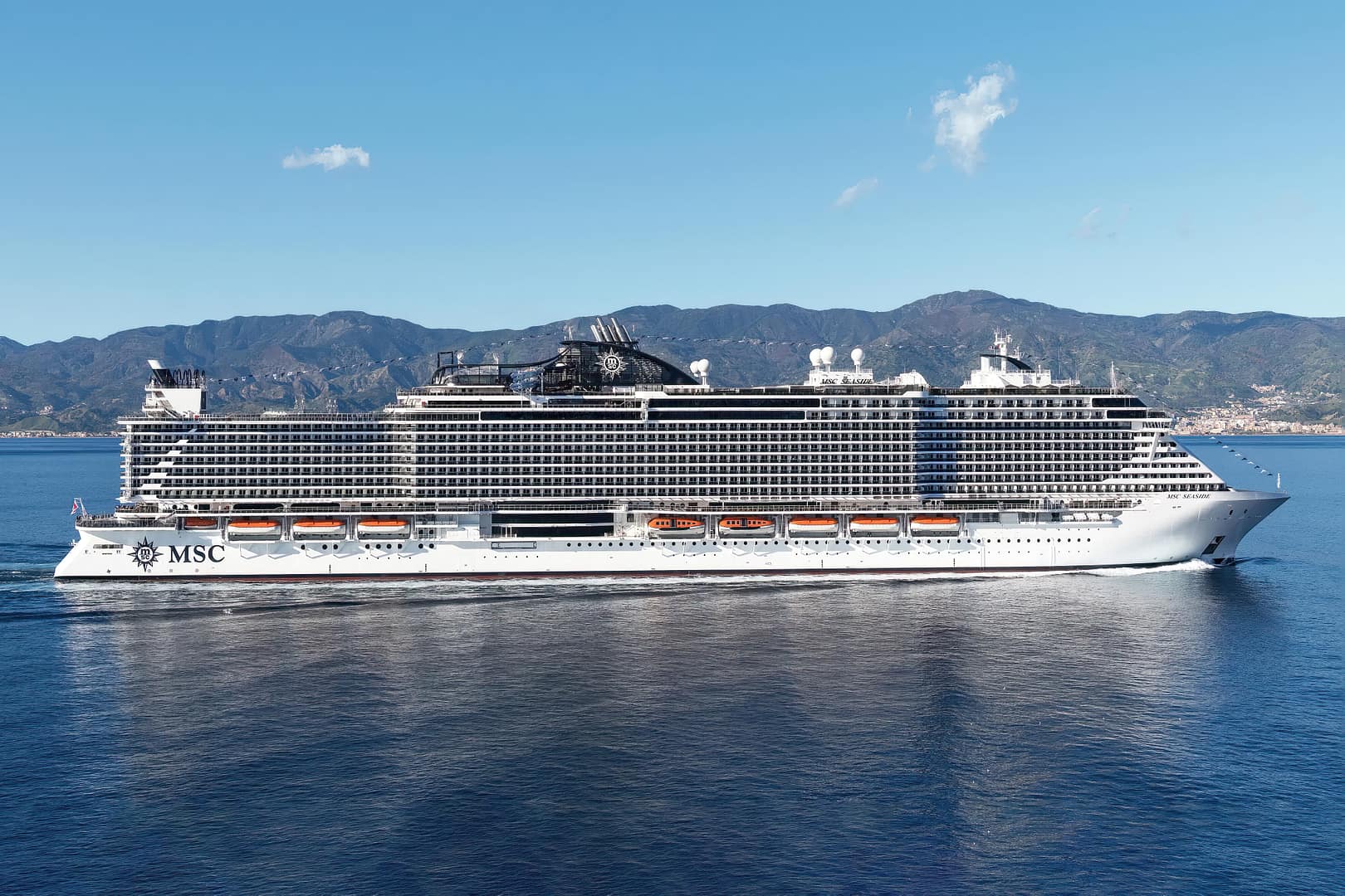 A large MSC Seaside cruise ship floating on a calm sea with a mountainous coastline in the background under a clear blue sky