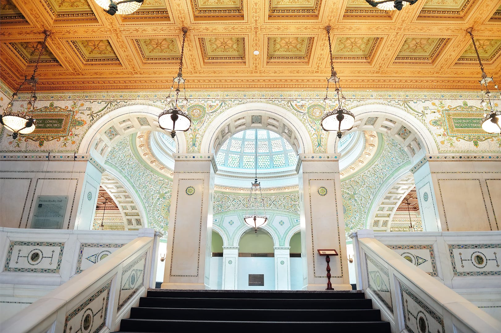 Elegant interior of the Chicago Cultural Center with ornate ceilings, intricate tile work, grand staircases, and hanging lamps, leading to a large arched window