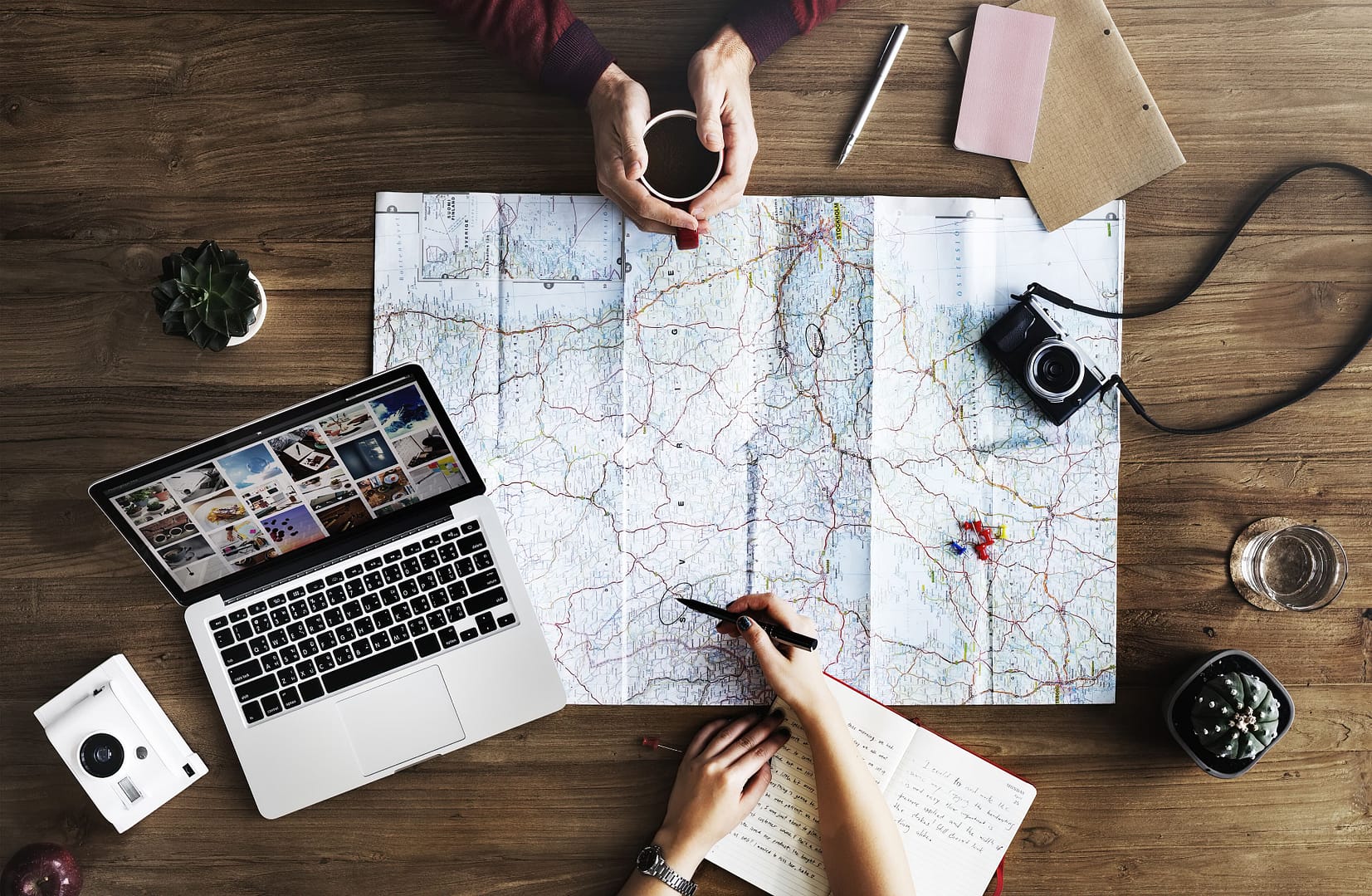 Two people conducting travel research using a map on a wooden table, surrounded by a laptop, camera, notebook, and travel accessories