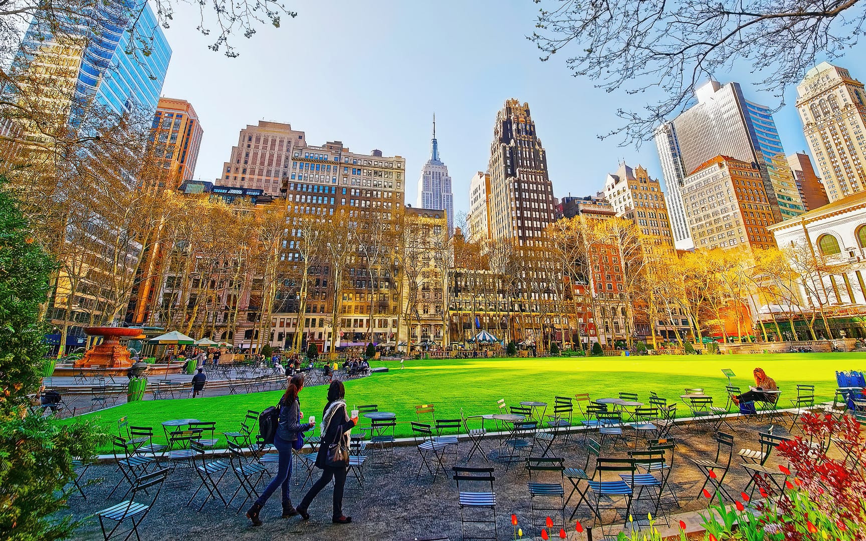Bryant Park in New York City on a sunny day, featuring a lawn surrounded by skyscrapers, empty chairs and tables in the foreground, and people walking
