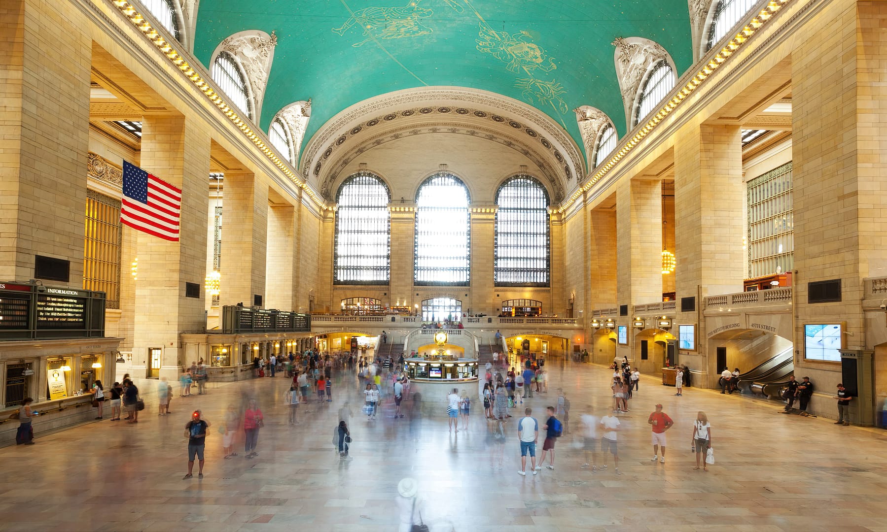 Interior of Grand Central Terminal in New York City, showing busy main concourse with large windows and American flags