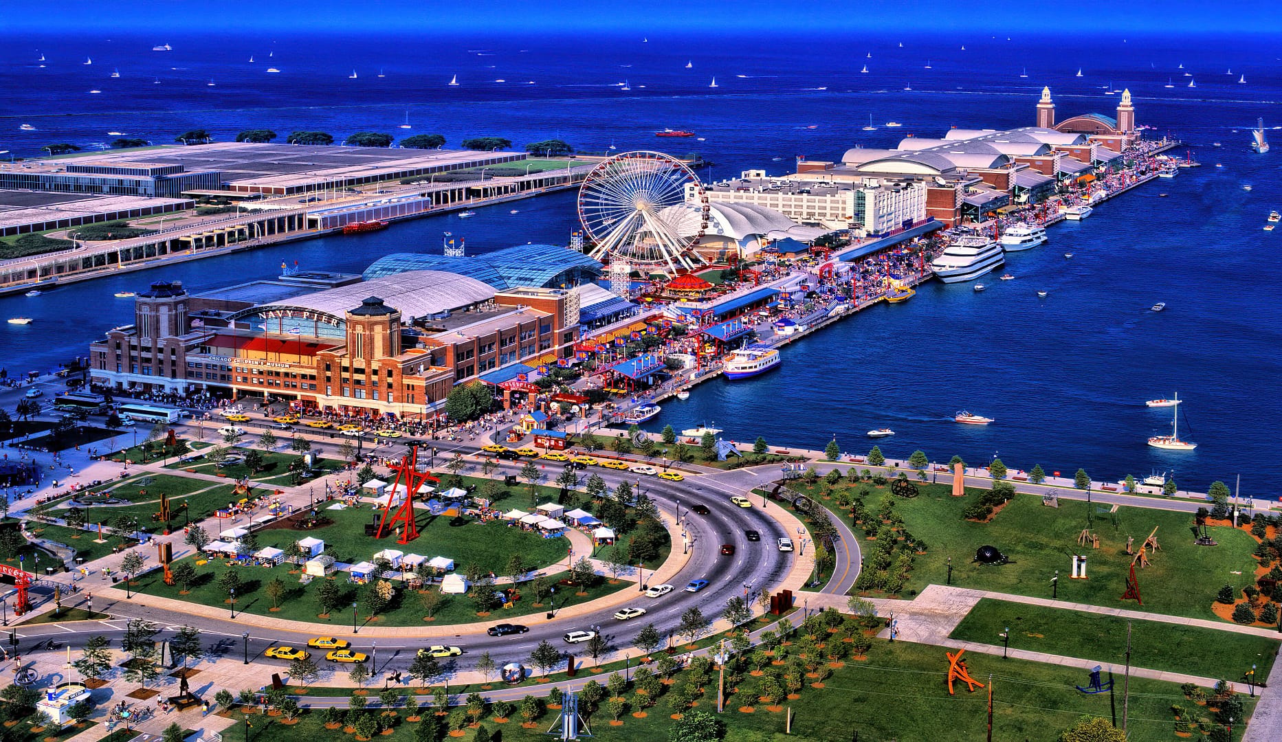 Aerial view of Navy Pier, a bustling waterfront district with a ferris wheel, large buildings, and a busy street, surrounded by clear blue water