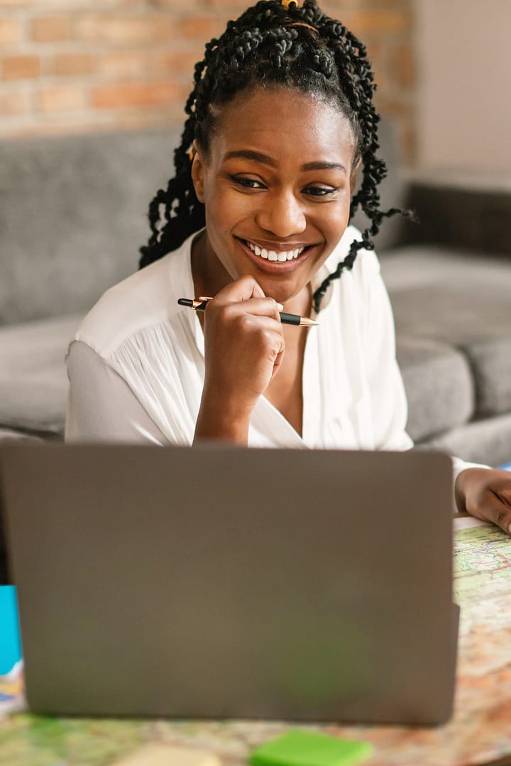 Happy black woman planning vacation with map and laptop, choosing best travel routes for future journey and preparing for trip, sitting at home
