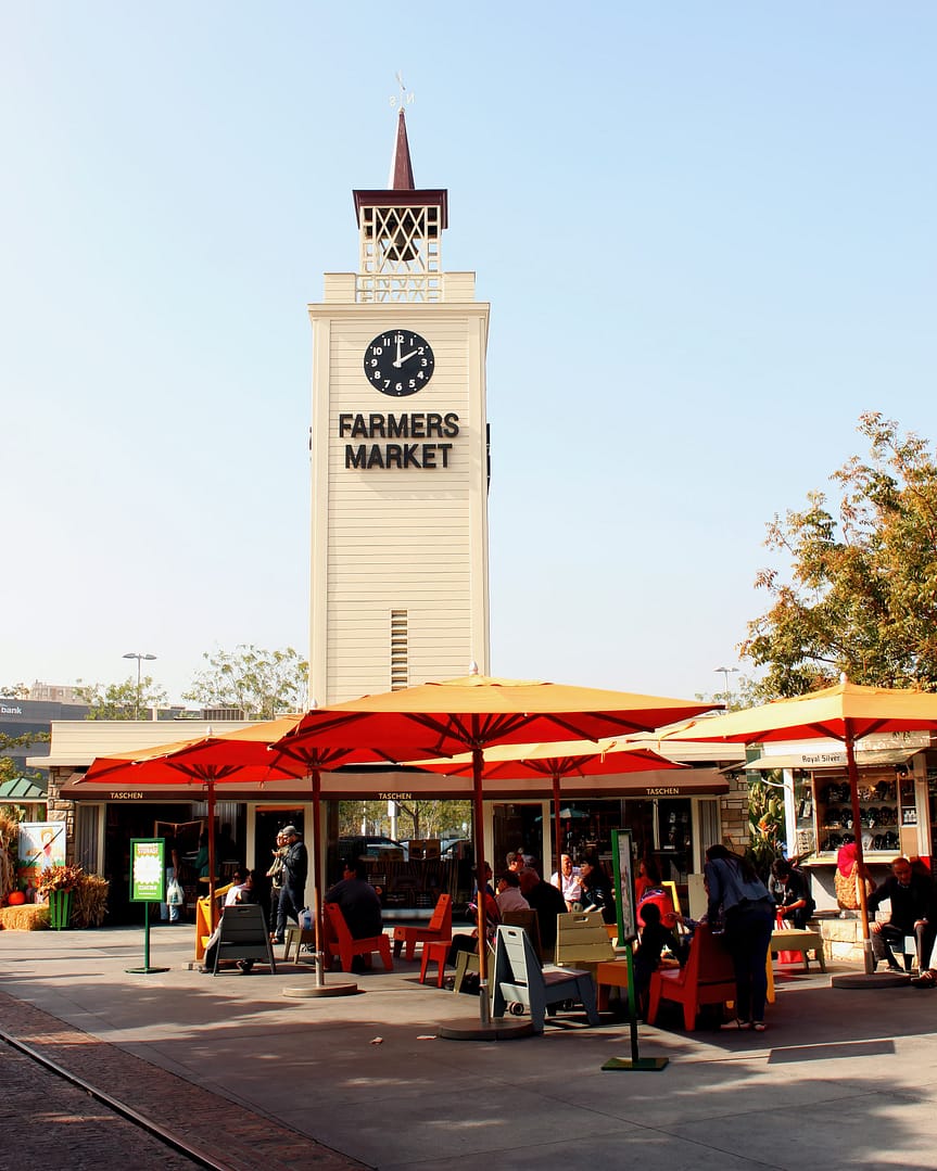 A large white clock tower exterior and people outside of The Original Farmers Market