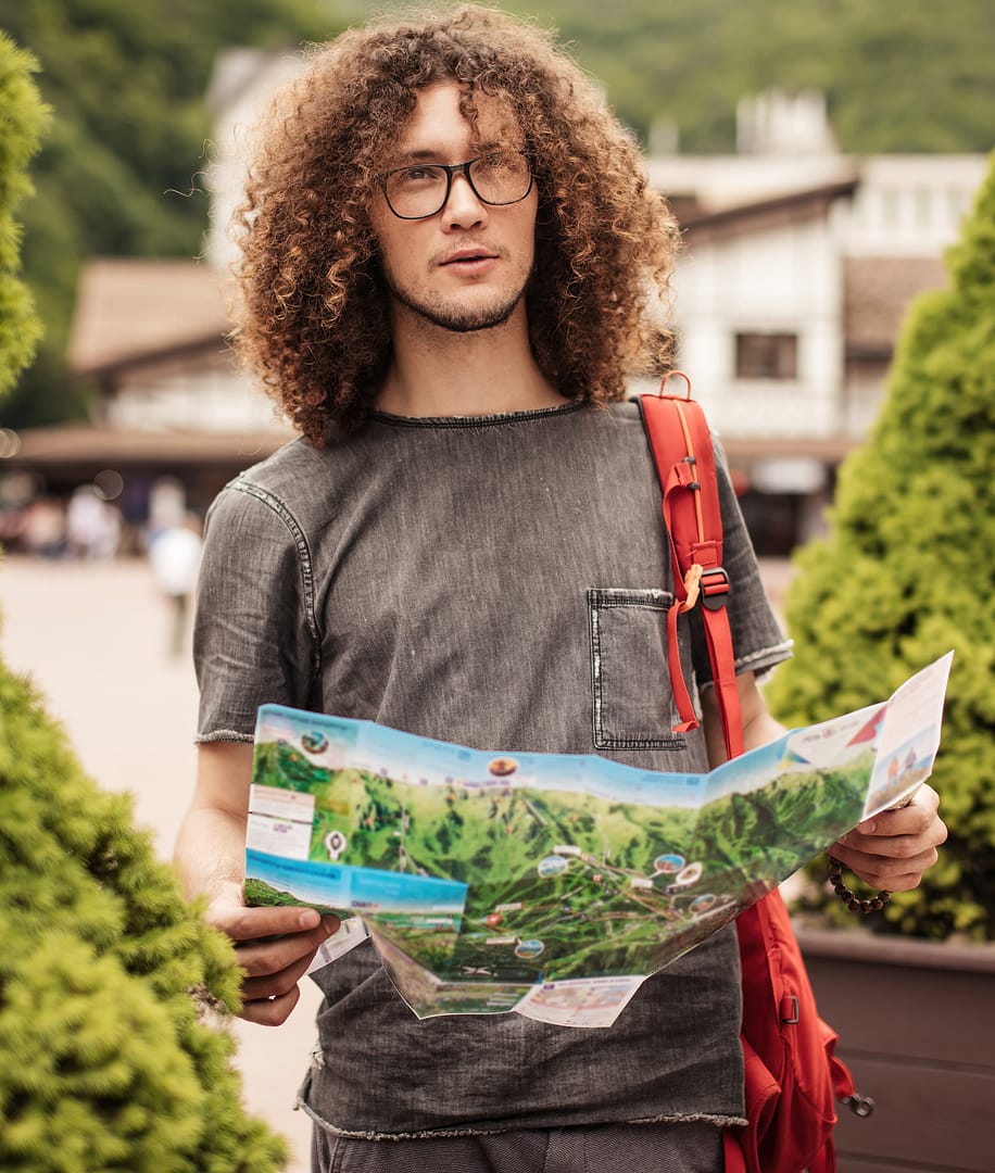Portrait of young curly-haired man traveler with glasses wearing a backpack and holding a map is choosing a location