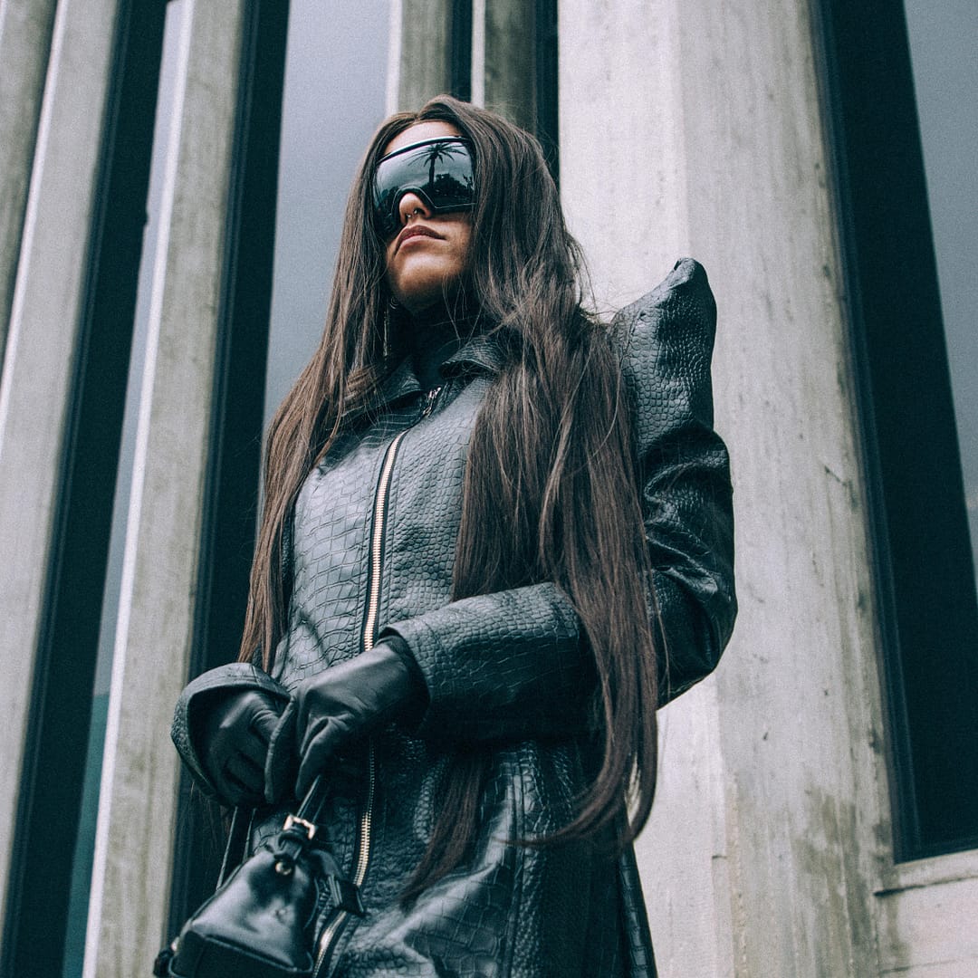 Woman in a long black coat and sleek sunglasses stands poised before a building with tall, reflective windows