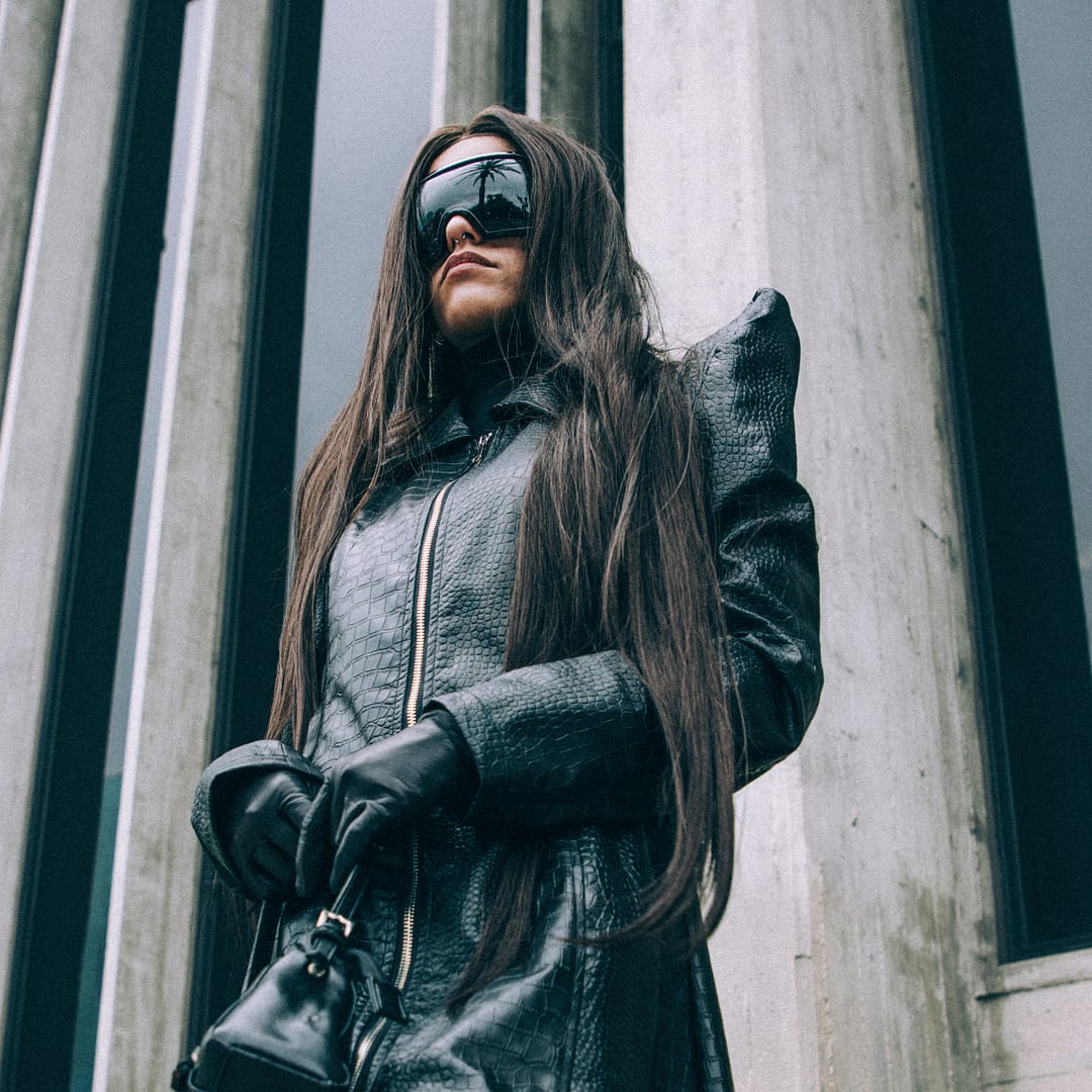 Woman in a long black coat and sleek sunglasses stands poised before a building with tall, reflective windows