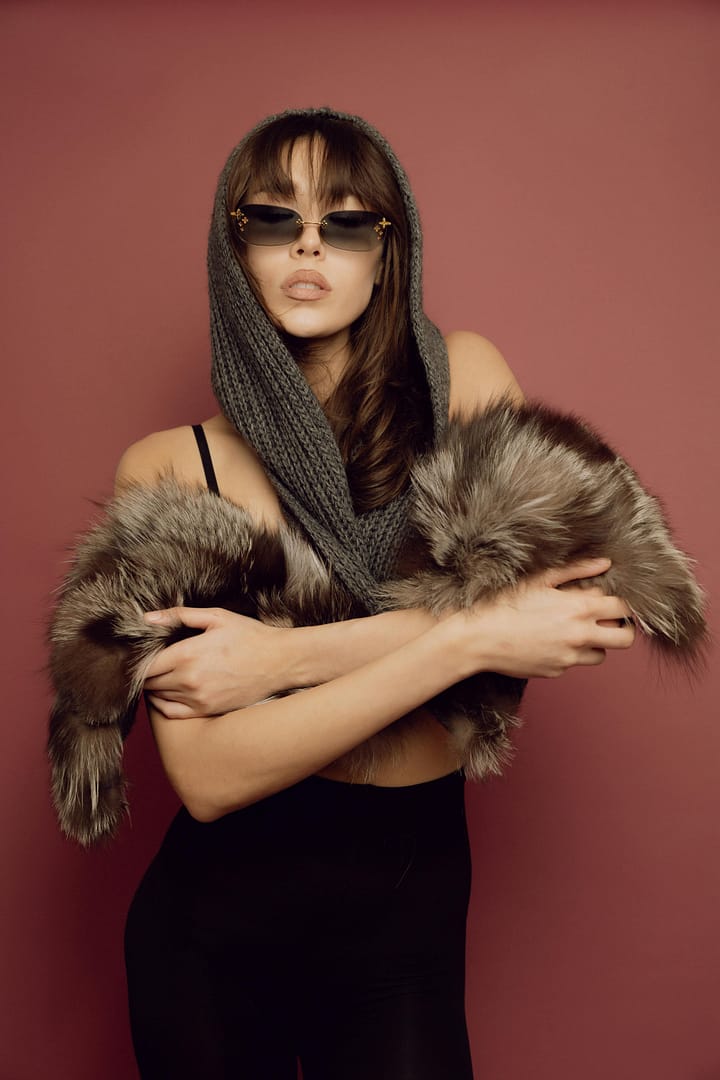 Woman wearing sunglasses and a knitted hood poses against a reddish background, draped in a faux fur garment