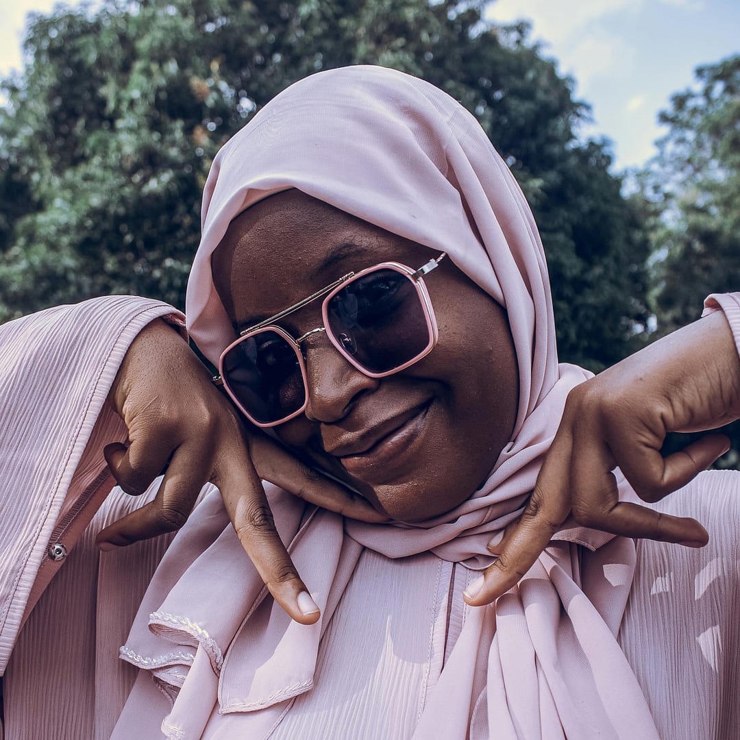 Black woman wearing a pink hijab and stylish sunglasses is smiling warmly while making a friendly hand gesture outdoors