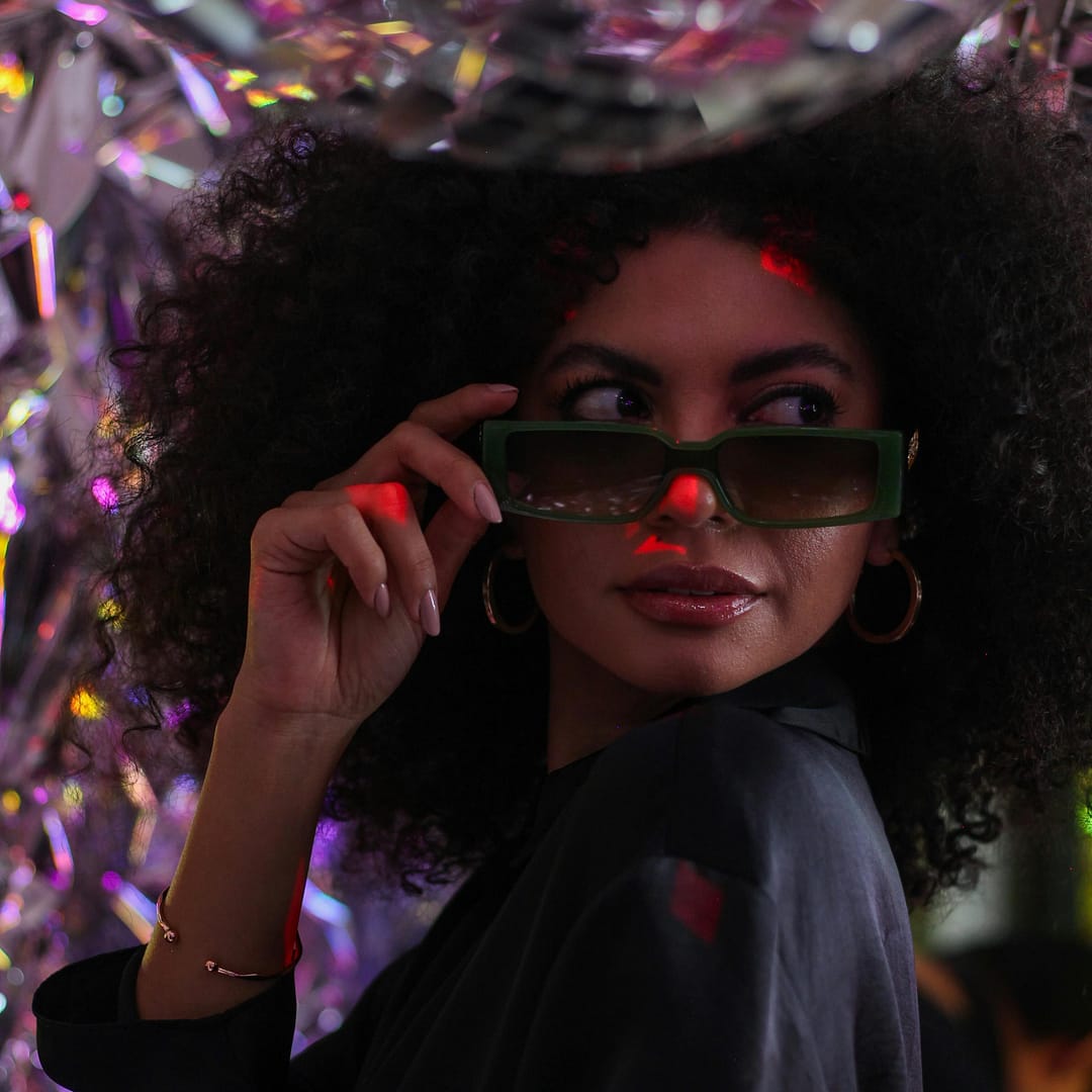Woman with curly hair dons sleek sunglasses and a dark dress, standing amidst reflective decorations illuminated by vibrant lighting