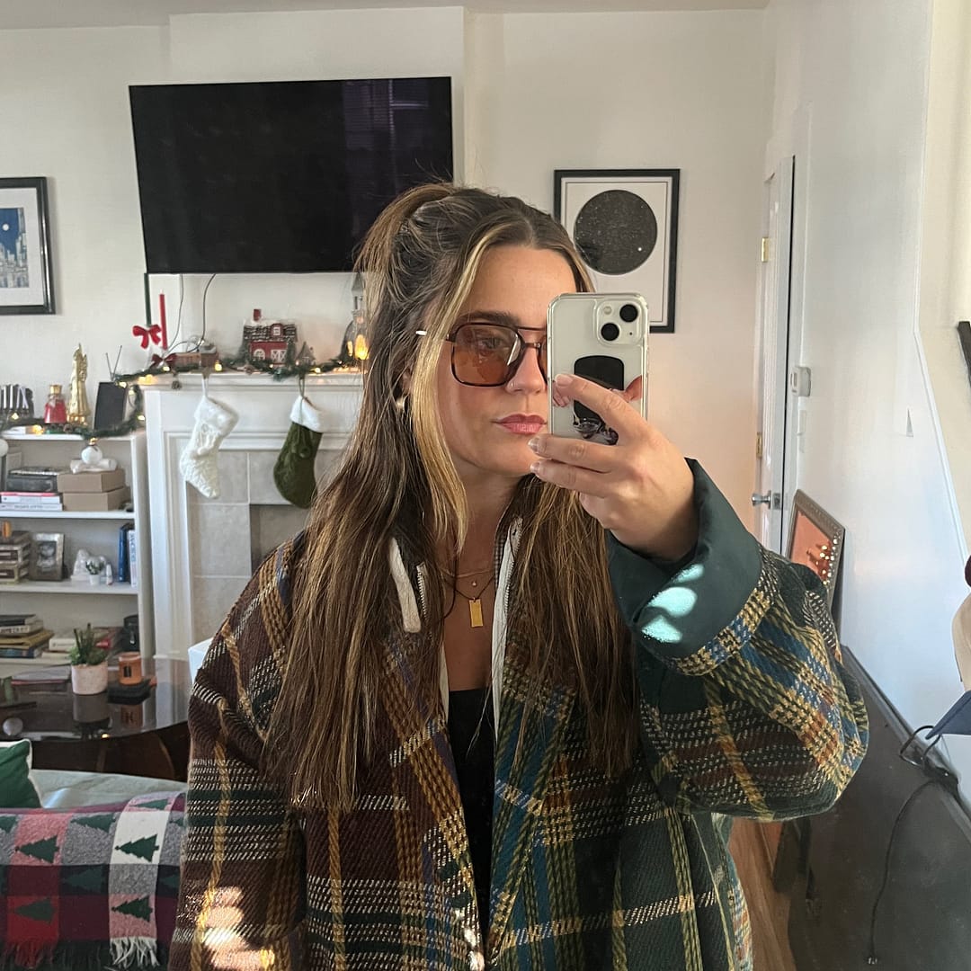 Woman with heart-shaped face in stylish sunglasses and a plaid coat snaps a mirror selfie in a living room adorned with chic wall art, a sleek TV, and minimalist shelves